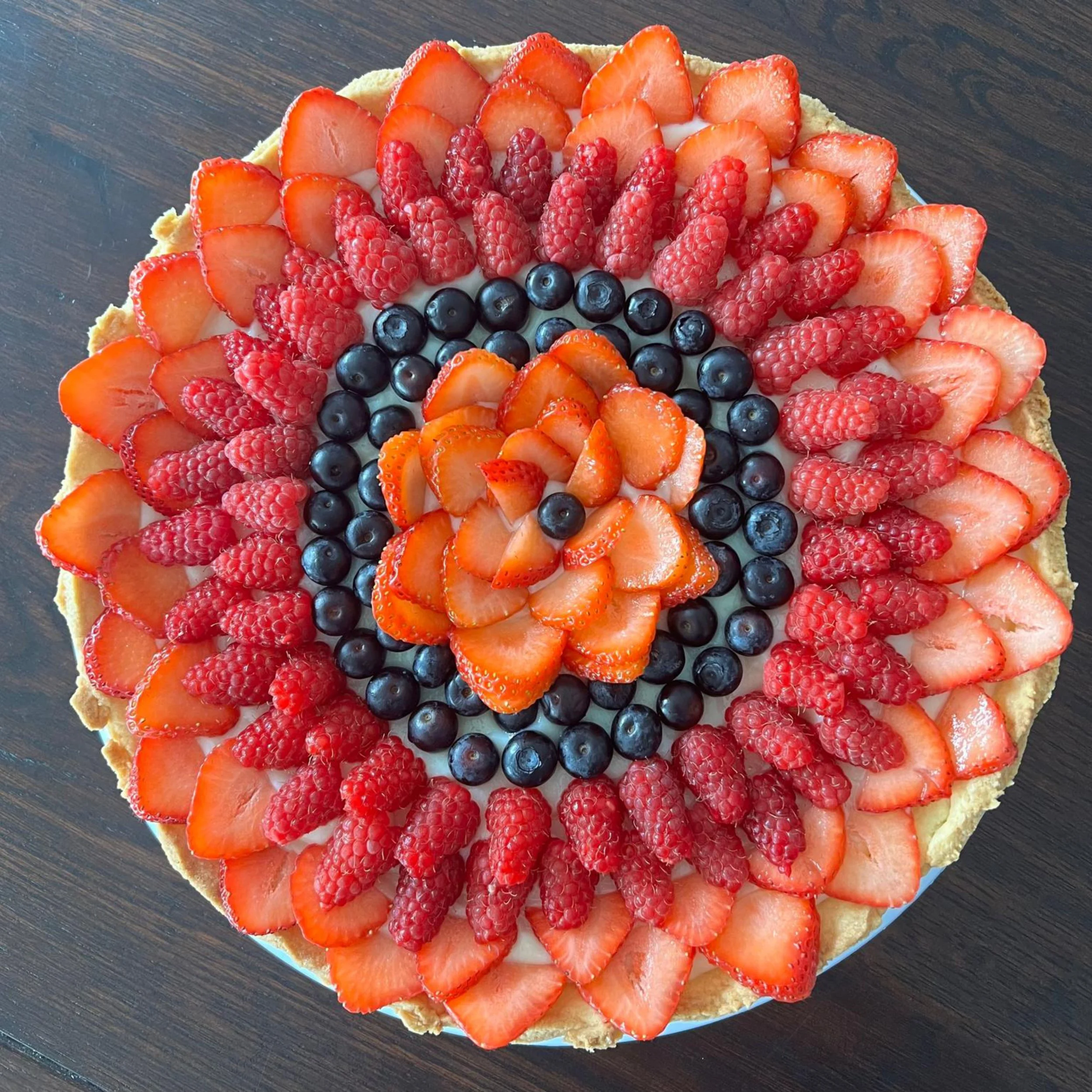 A round fruit tart topped with sliced strawberries, raspberries, and blueberries arranged in concentric circles.