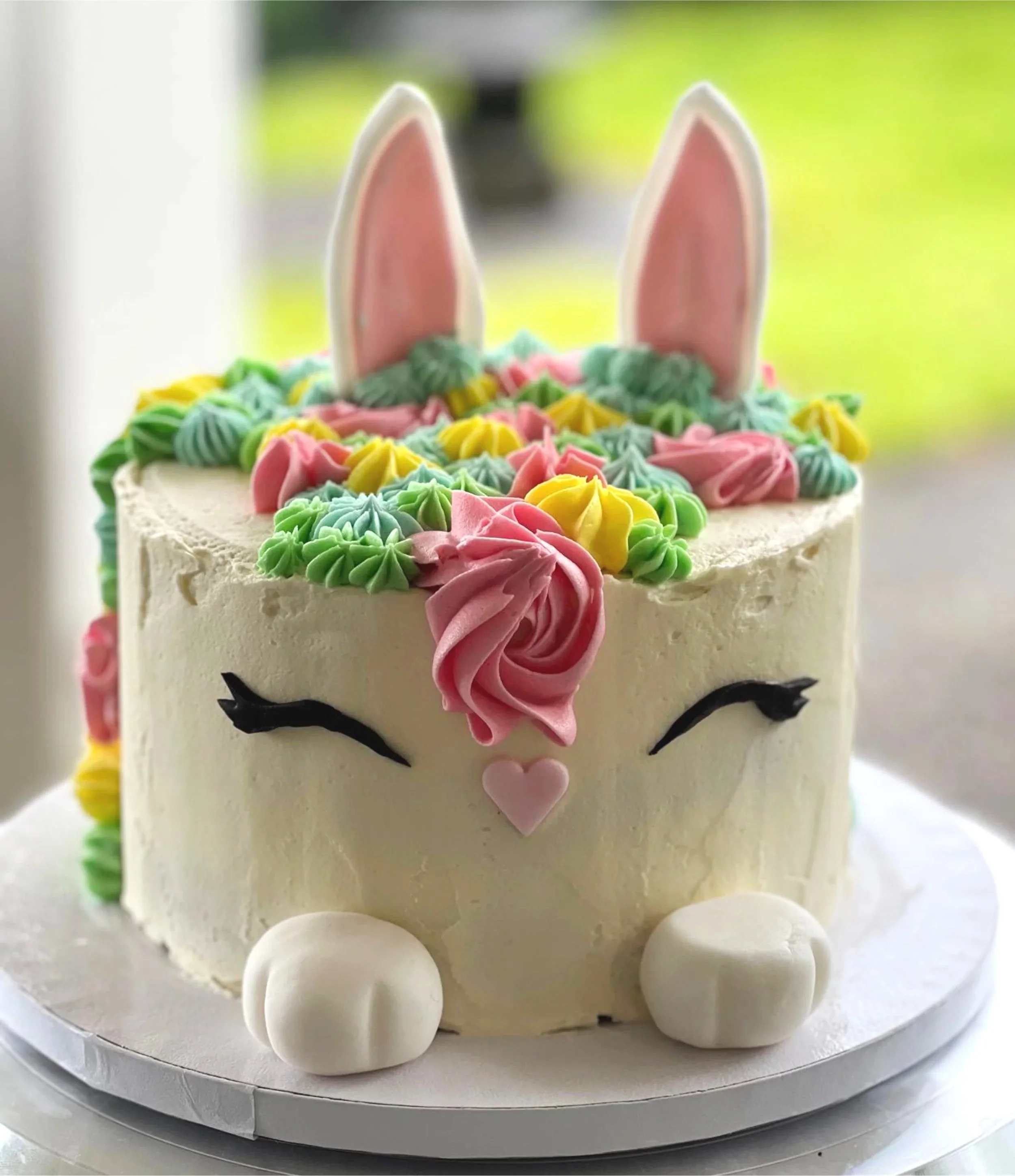 Unicorn-themed birthday cake decorated with pastel-colored frosting, featuring closed eyes with long eyelashes, pink and white ears, a pink heart-shaped nose, and two white spherical decorations resembling hooves.