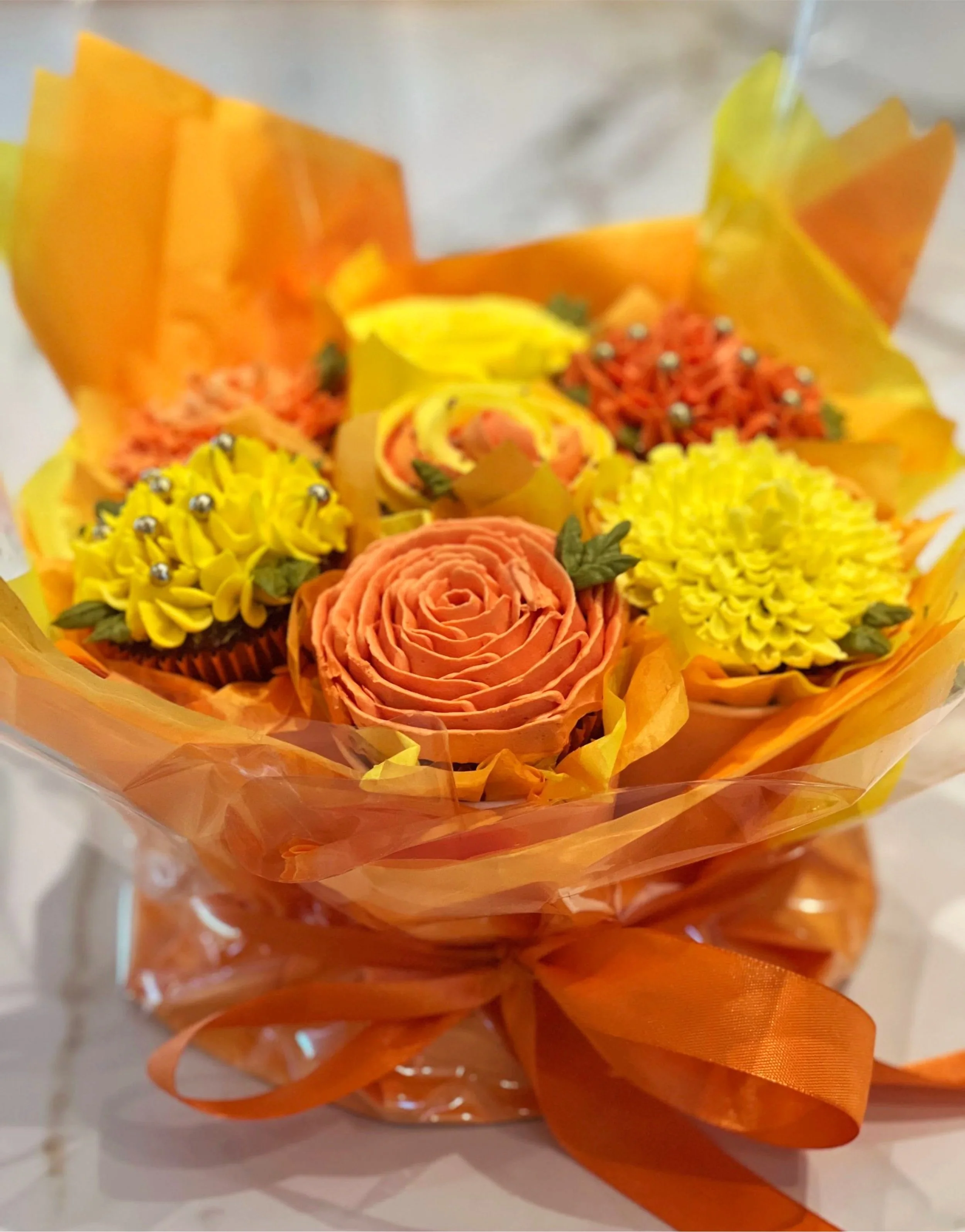 A bouquet of cupcakes decorated to look like flowers in orange, yellow, and peach colors, wrapped in orange cellophane with an orange ribbon.