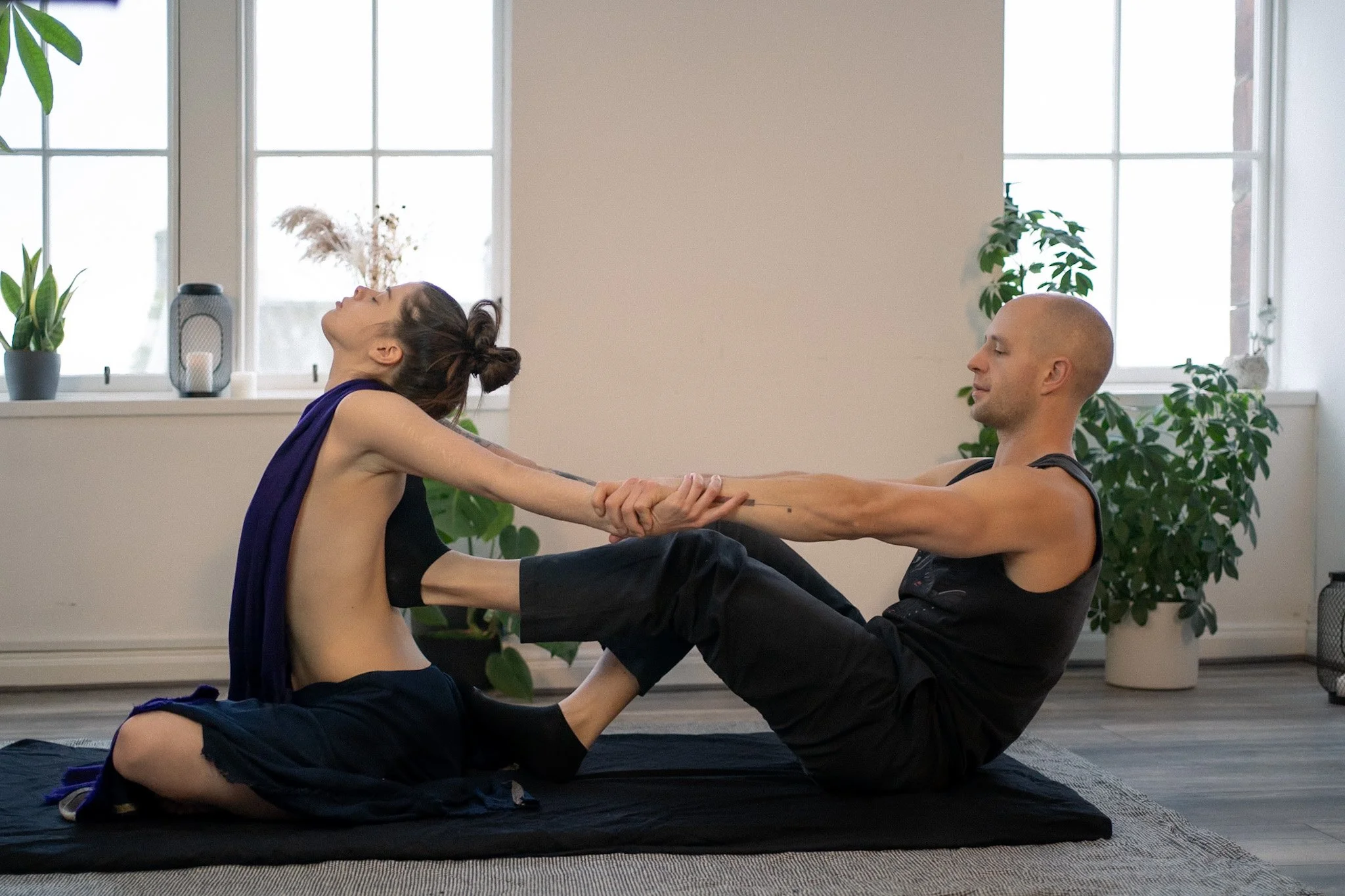 An Ayurvedic Yoga Massage therapist giving a treatment to a woman, with the woman leaning back and the therapist supporting her while sitting on the floor in front of him, holding her arms and assisting her.