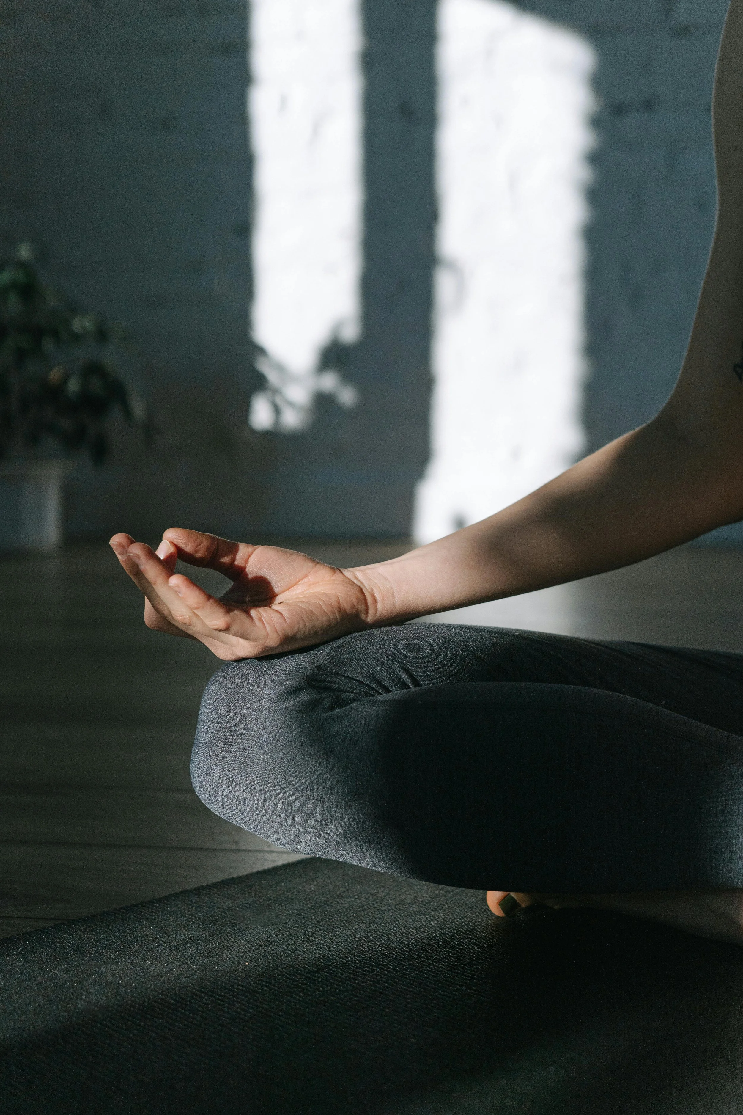 Person seated on a black yoga mat with a toned arm extended, practicing meditation or yoga in a dimly lit room with sunlight casting shadows on the wall.