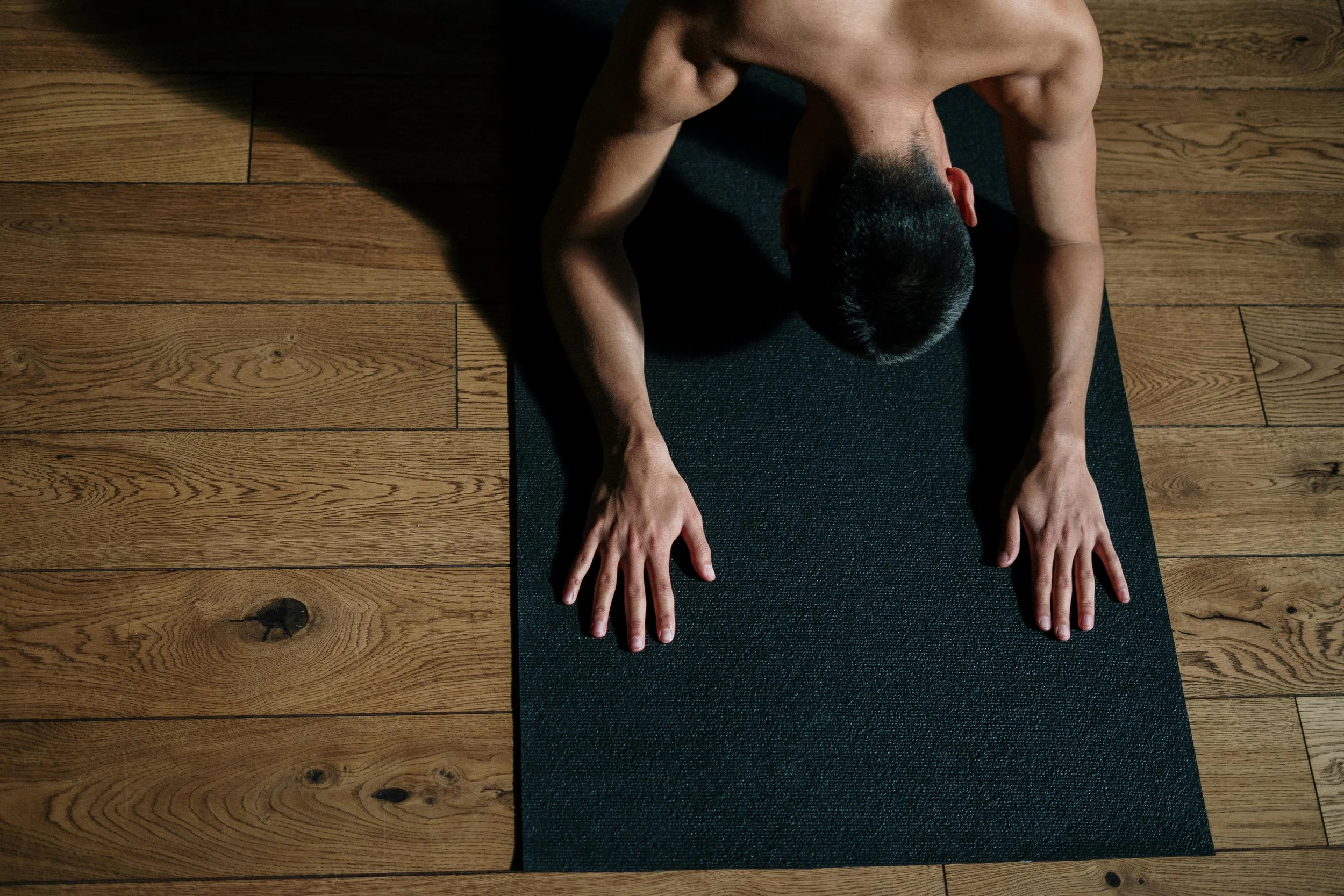 A man practicing yoga on a dark mat on a wooden floor, seen from above in a forward bend pose.