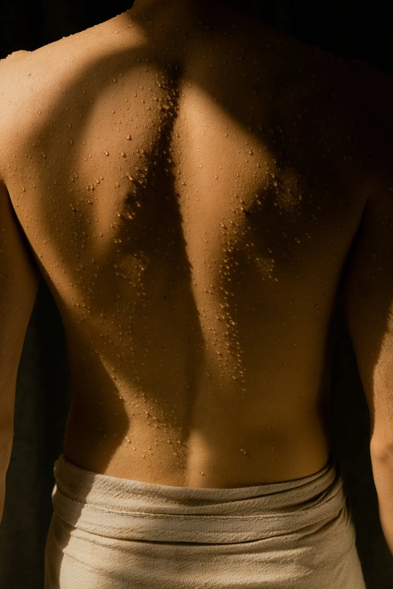 Back of a shirtless man with moist skin and water droplets, wearing light-colored pants, with a shadow cast on his back from side lighting.