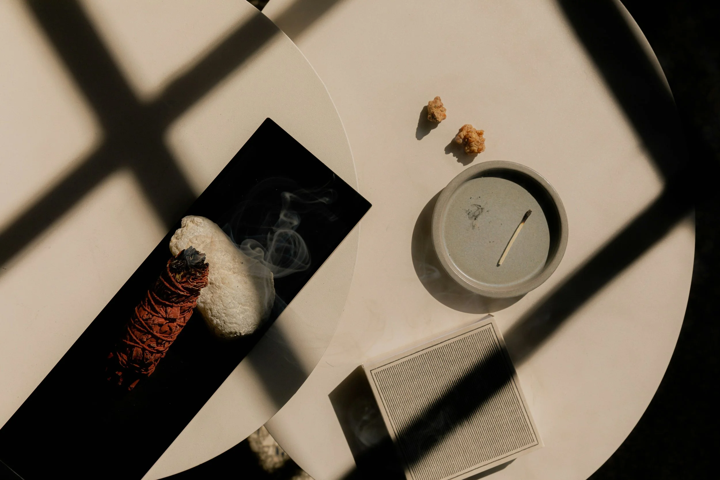 A beige round table with a candle, ceramic mug, small rocks, and a rectangular black dish containing a curling incense stick, a rock, and smoke.