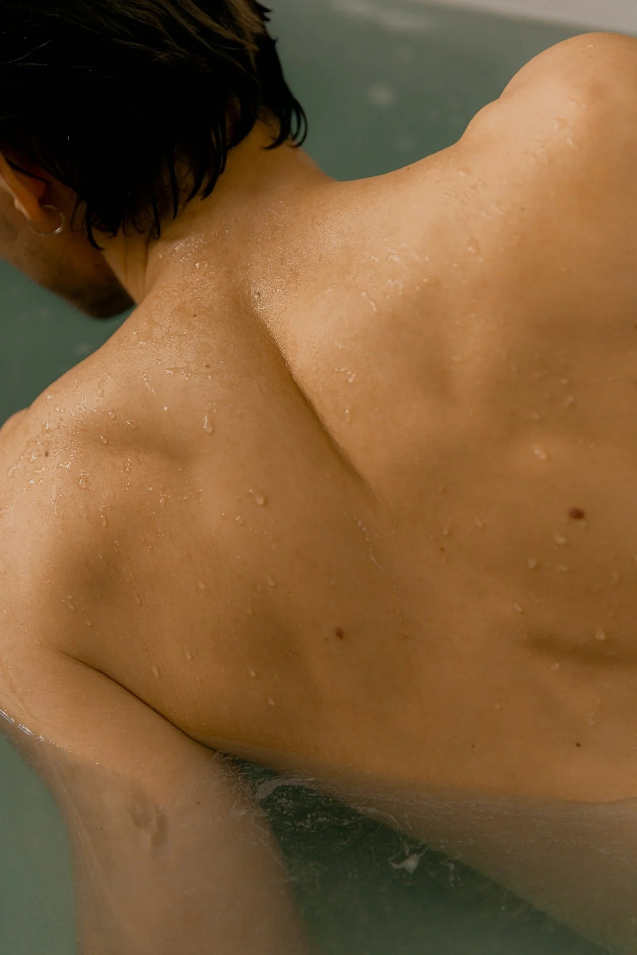Close-up of a person's upper back, shoulder, and neck in a shower with water droplets on the skin.