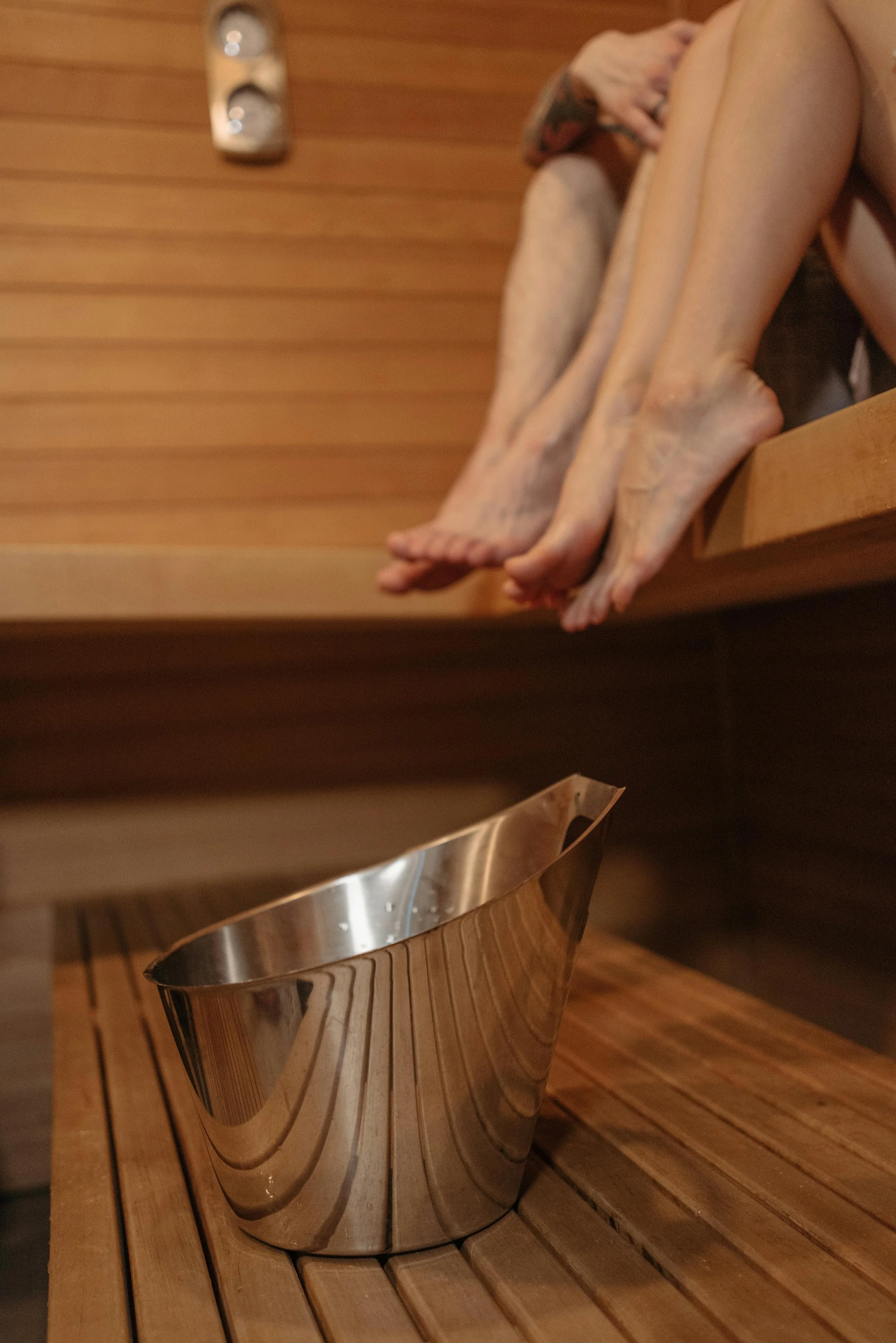 View inside a sauna with a metal bucket on a wooden bench and a person sitting with their legs crossed in the background.