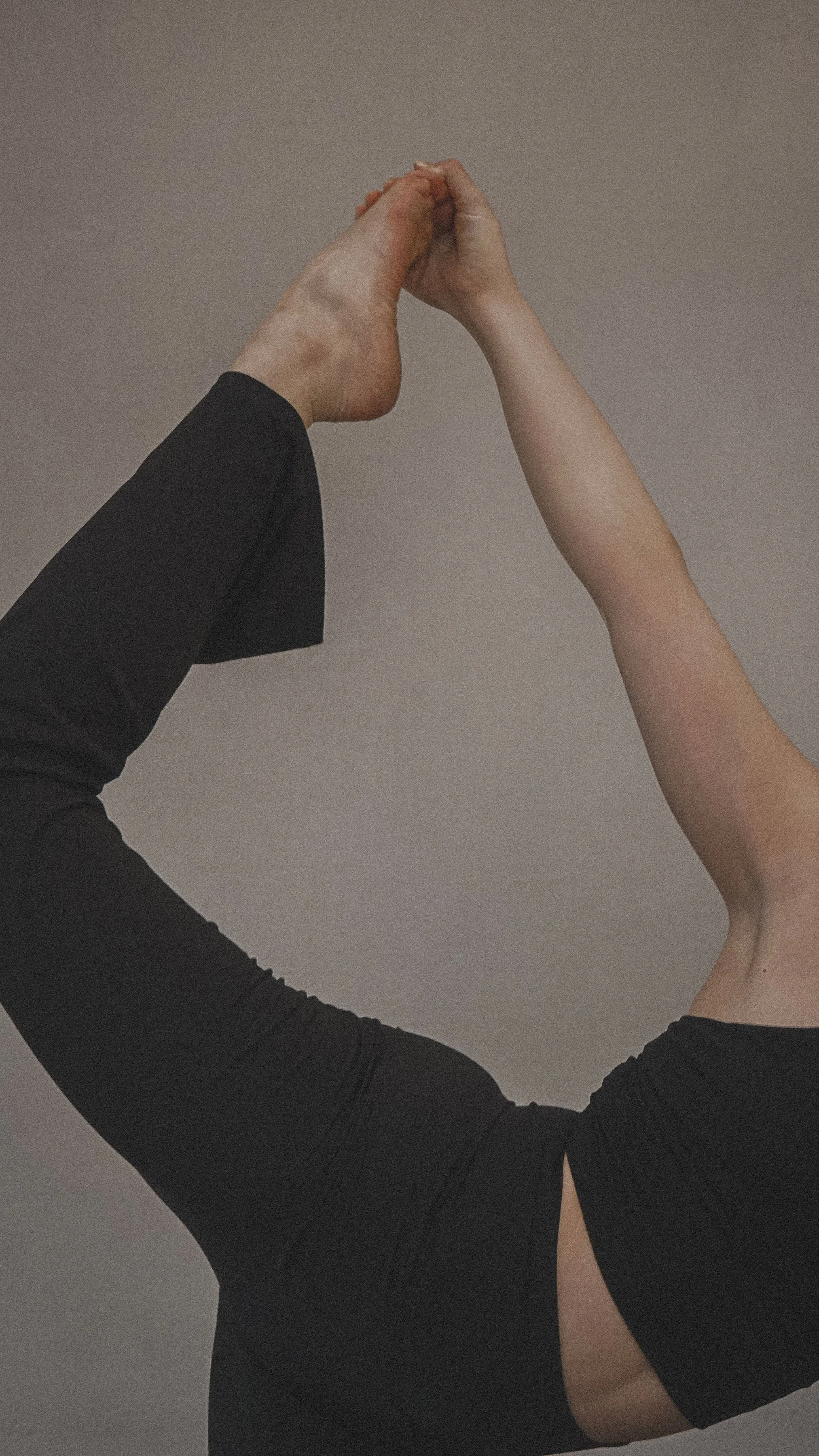 Close-up of a person practicing yoga, holding their foot behind their back with one hand, wearing black athletic clothing, against a plain background.