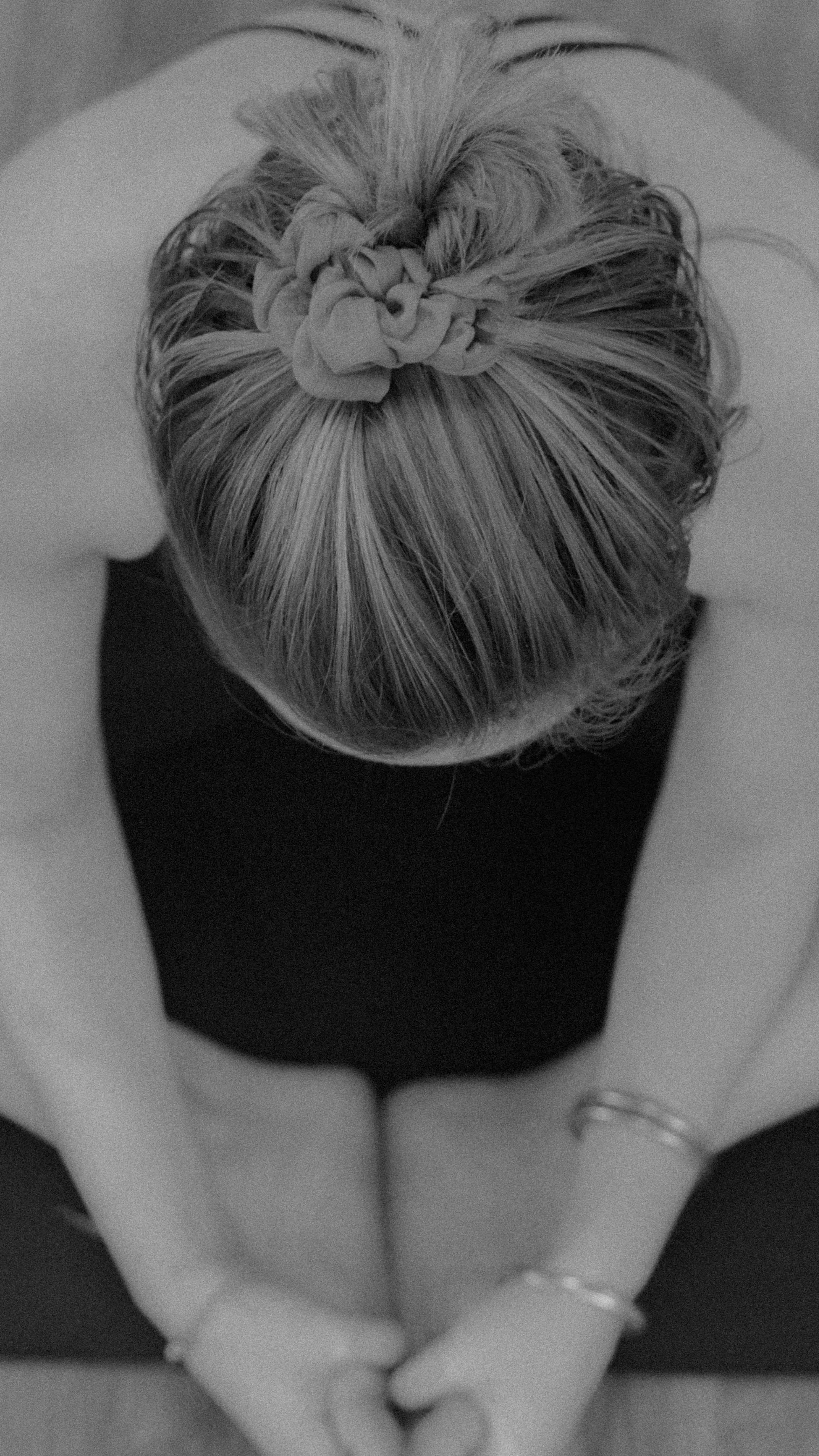 Top-down view of a woman with blond hair in a bun with a scrunchie, wearing a black sleeveless top, hands clasped in front of her, and wearing rings on her fingers.