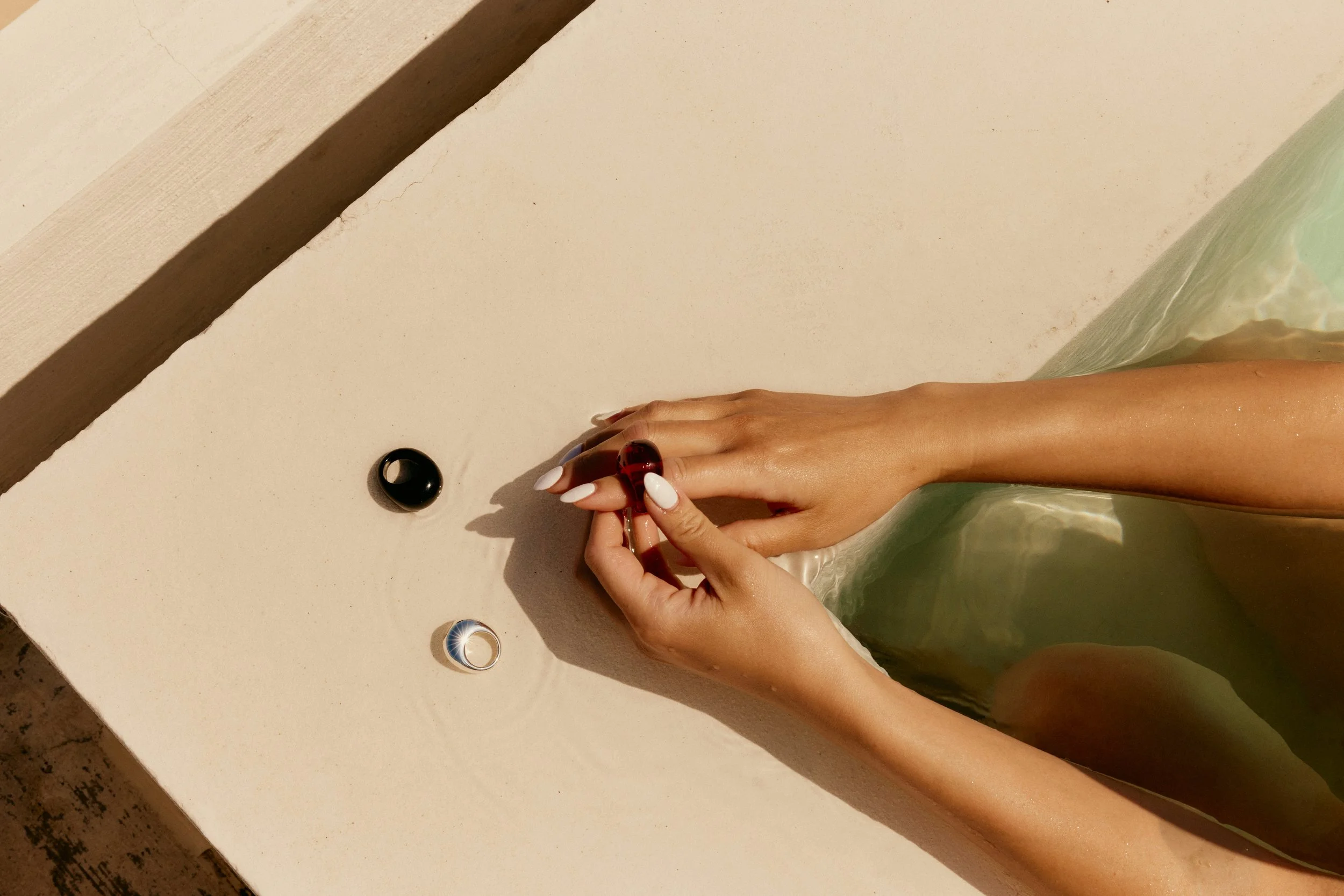 A person with manicured white nails is holding a small red object, partially submerged in a bathtub. Two other small objects are on the edge of the bathtub, one black and one metallic.