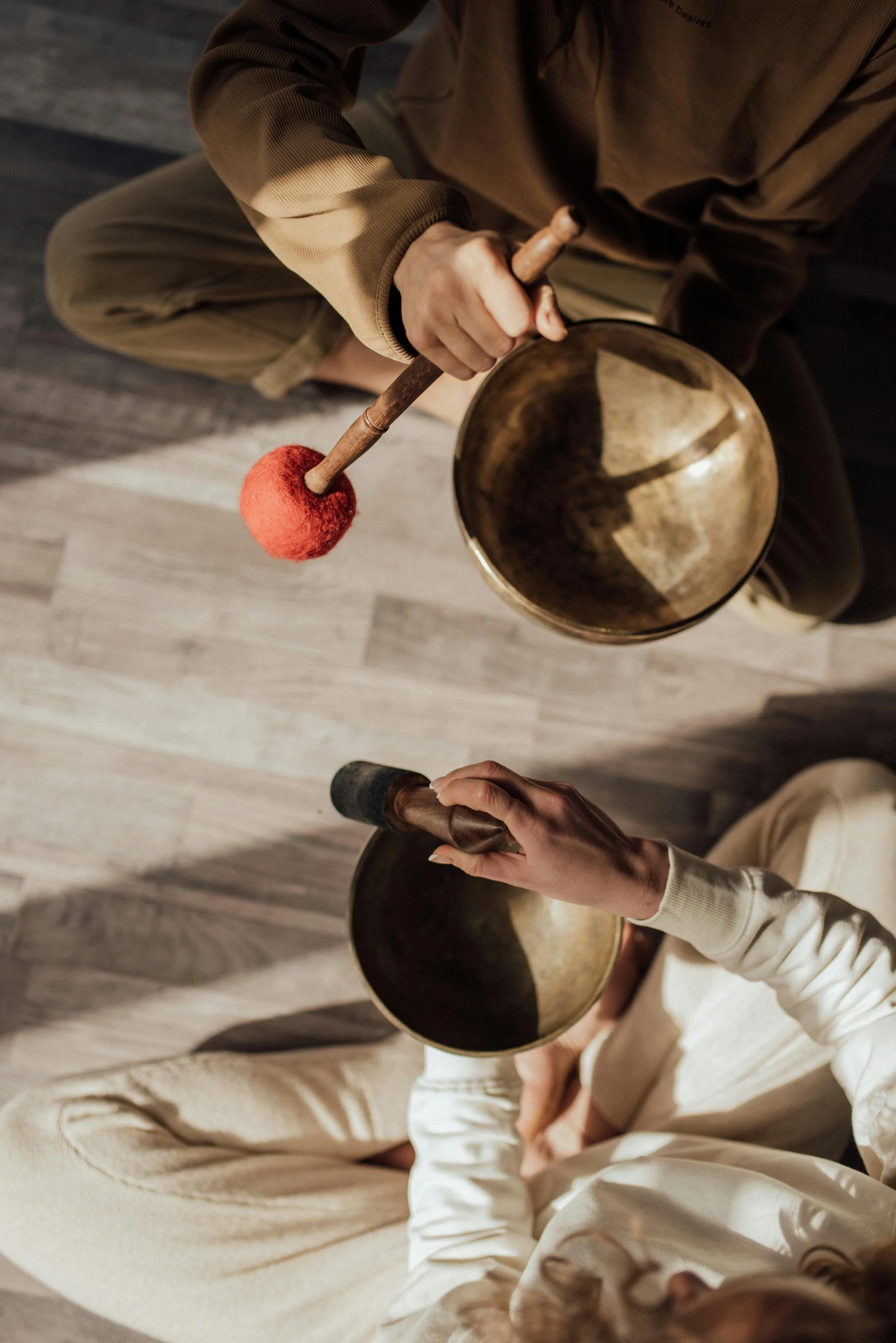 Two people are sitting on the floor playing singing bowls with mallets. One person is holding a red felt mallet, and the other is holding a black mallet. The image is shot from above, showing the top view of their hands and the bowls.