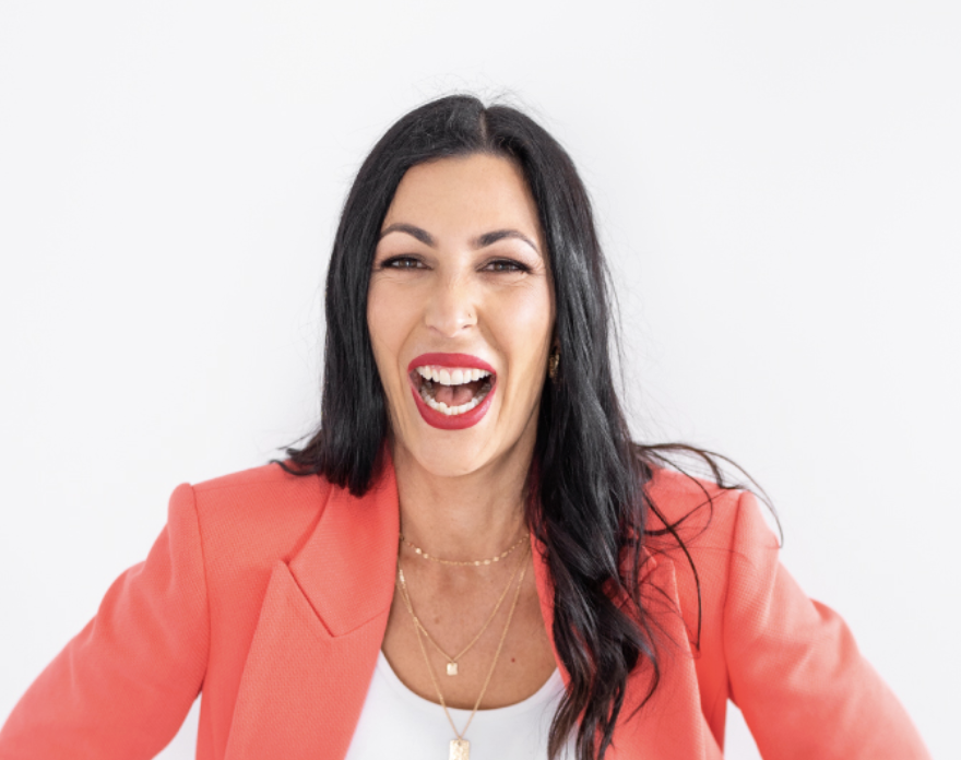 Woman with black hair, red lipstick, wearing a coral blazer and layered necklaces, smiling and laughing against a plain white background.