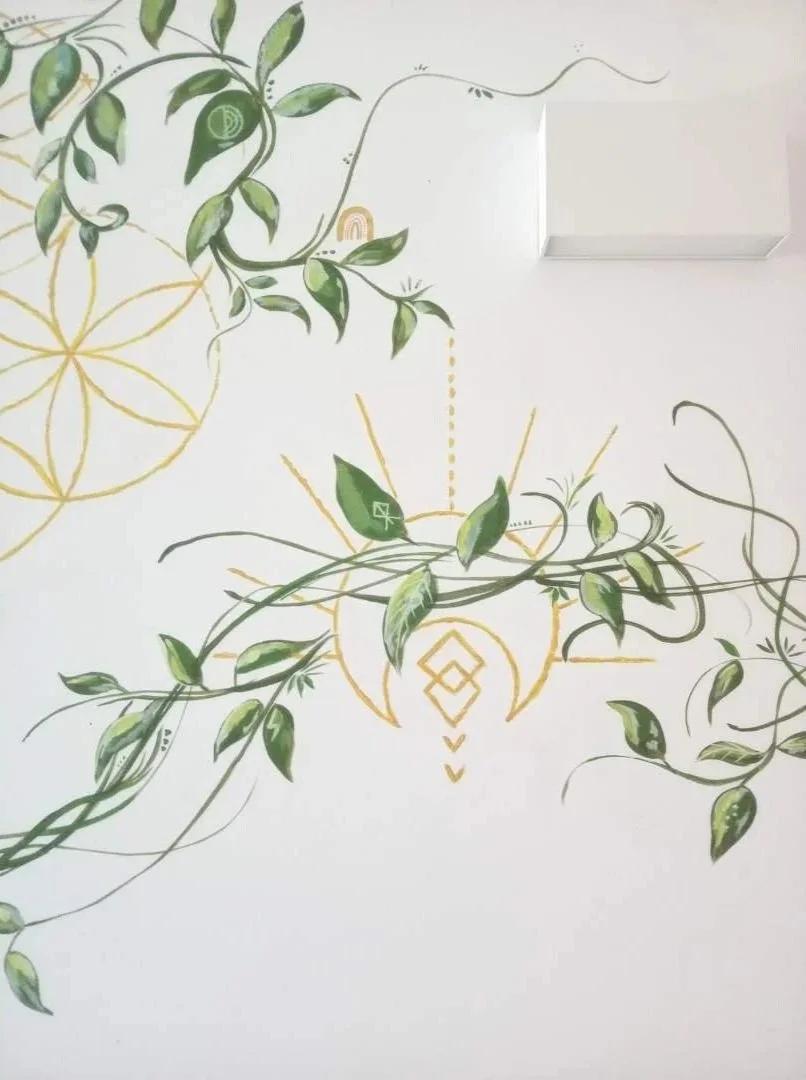 Wall mural with green leafy vines and geometric gold patterns on a white wall, including an abstract hanging plant and a white box mounted on the wall.
