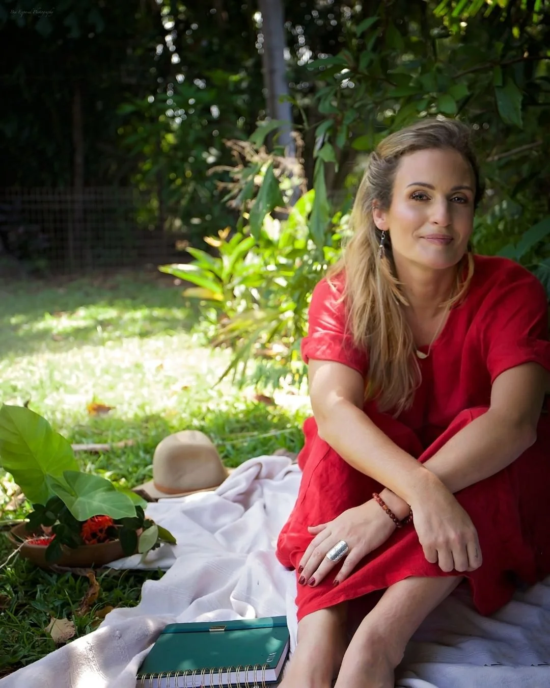 A woman with long blonde hair and wearing a red dress sits on a white blanket in a lush garden, smiling softly at the camera. There is a hat, a bowl of flowers, a notebook, and some greenery around her.