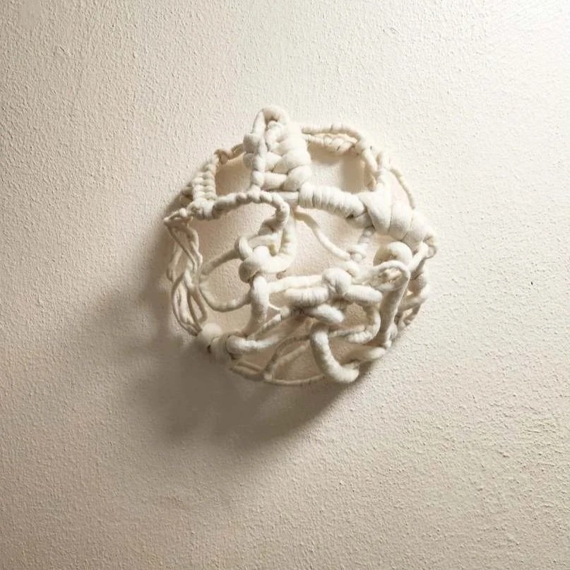 A white, abstract, tangled string art sculpture mounted on a plain white wall.