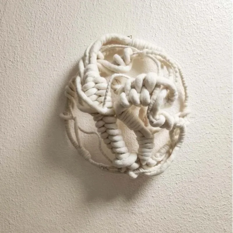 Abstract white textile sculpture mounted on a textured beige wall.