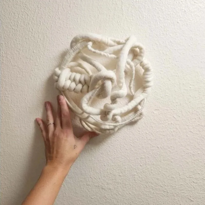 Hand reaching out to touch a piece of textured white wall art made from intertwined, soft, pipe cleaner-like material.