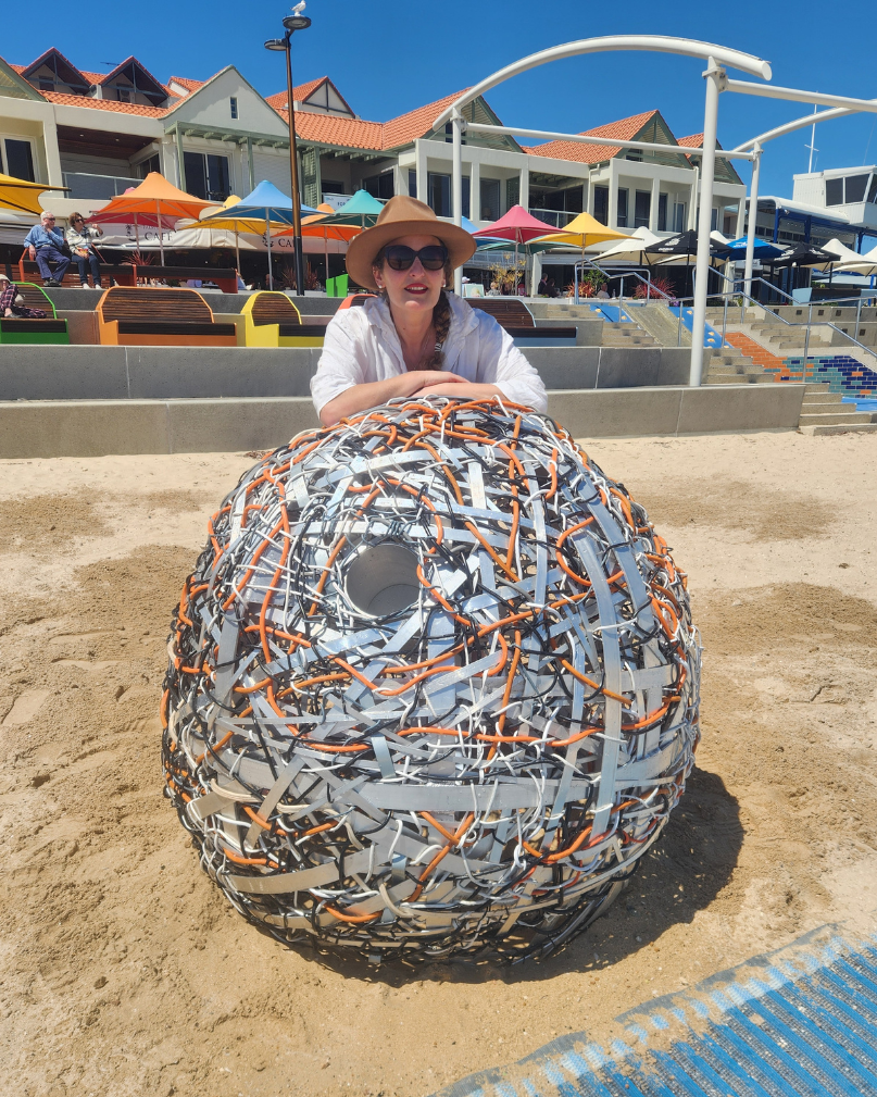 A woman wearing sunglasses and a hat sits behind a large, woven sculpture resembling a fish or sea creature made from metal scraps, on a sandy beach with colorful umbrellas and buildings in the background.