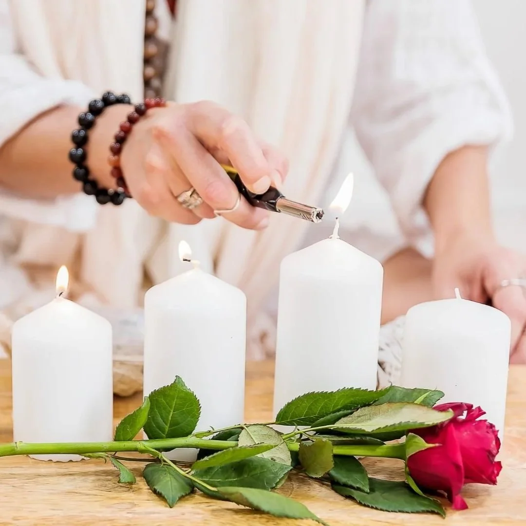 Person lighting a candle on a table with three white candles, a red rose, and green leaves.