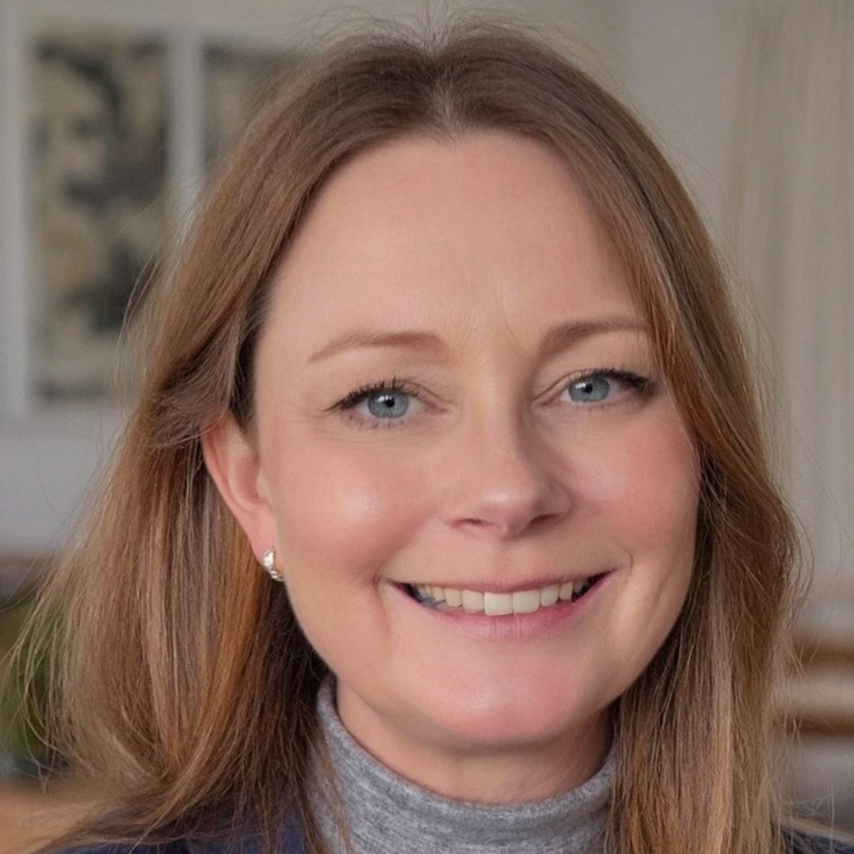 Close-up of a smiling woman with red hair, blue eyes, wearing pearl earrings and a gray turtleneck, indoors with neutral background.