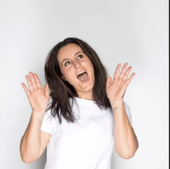 A woman with brown hair wearing a white t-shirt smiling and raising her hands near her face against a plain white background.