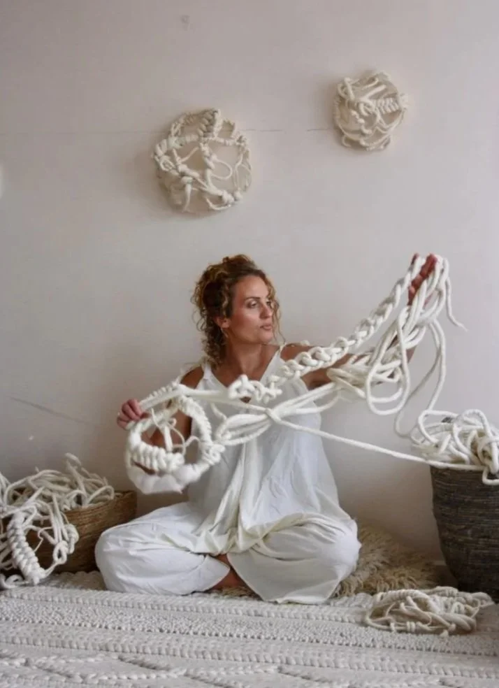 A woman in white sitting on a bed, holding a large, twisted, white rope sculpture. In the background, there are two wall hangings made of similar white ropes, and baskets filled with tangled white ropes on the floor.