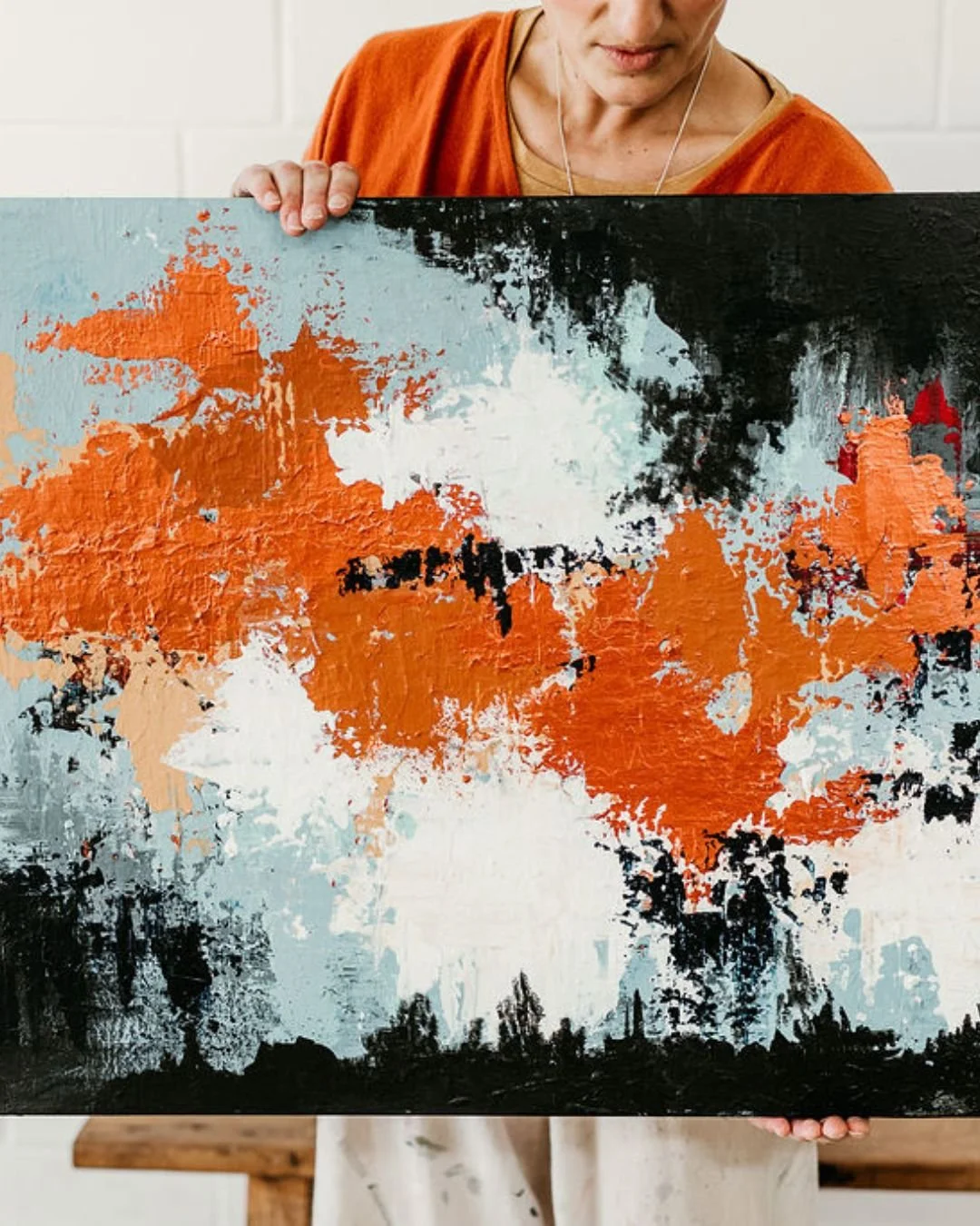 Person holding an abstract landscape painting with orange, black, white, and light blue colors.