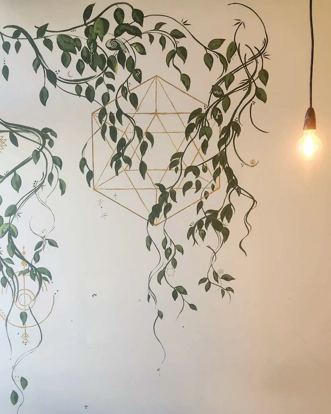 Wall art featuring a geometric design and green hanging vines with leaves, illuminated by a hanging light bulb to the right.