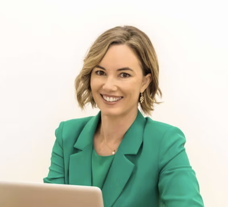 A smiling woman with short blonde hair, wearing a green blazer and earrings, sitting in front of a white background.