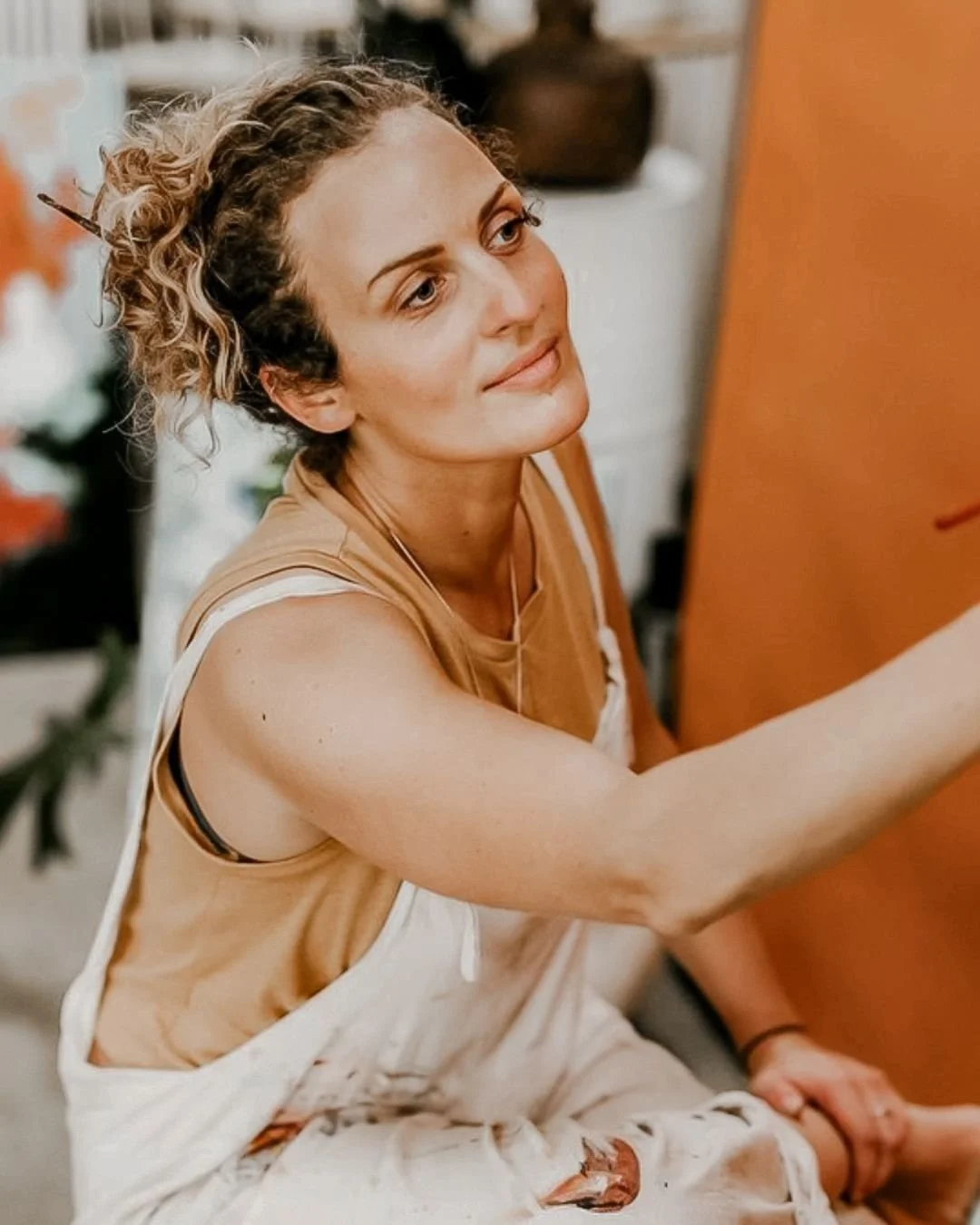 A woman with curly blonde hair in a messy bun, wearing a tan sleeveless top, sitting and smiling, painting on a canvas in an indoor setting.