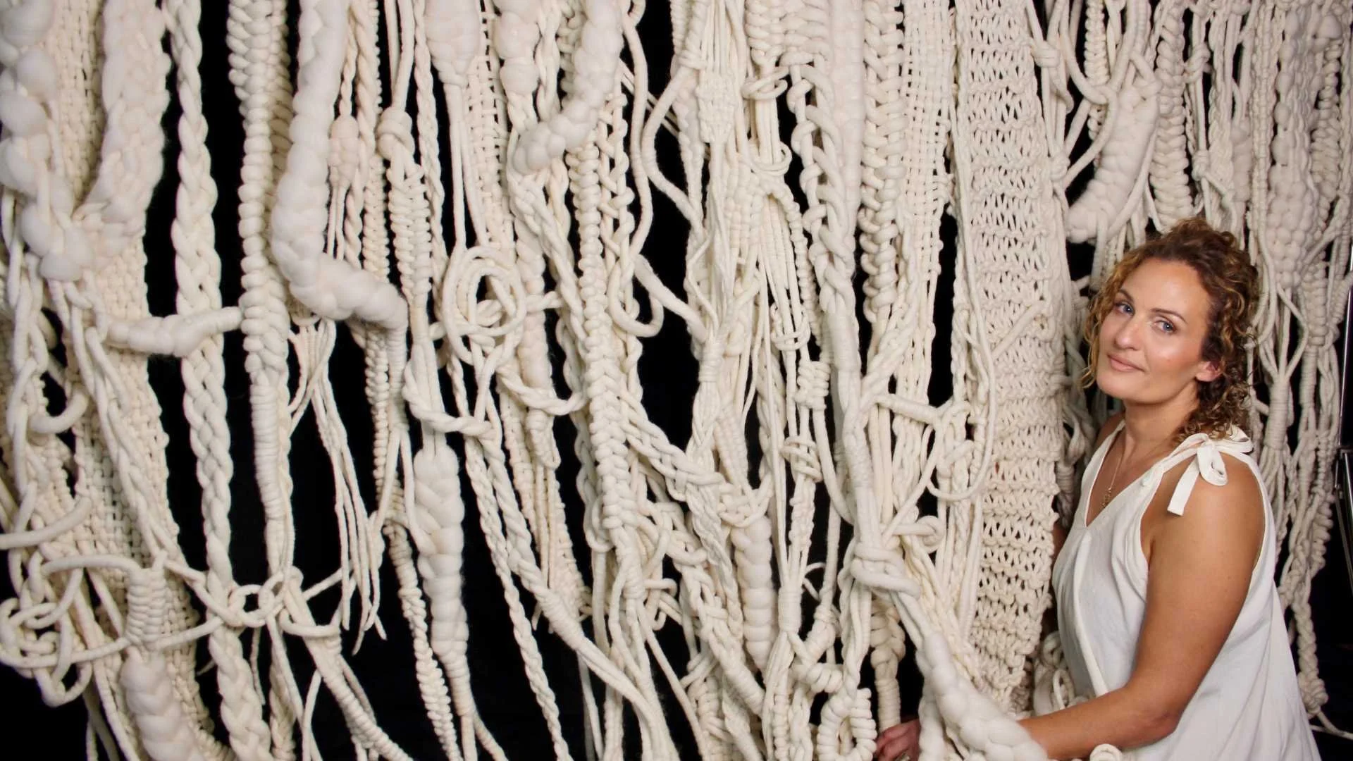 A woman with curly hair in a white sleeveless dress stands next to a large art installation made of white knotted and braided ropes against a black background.