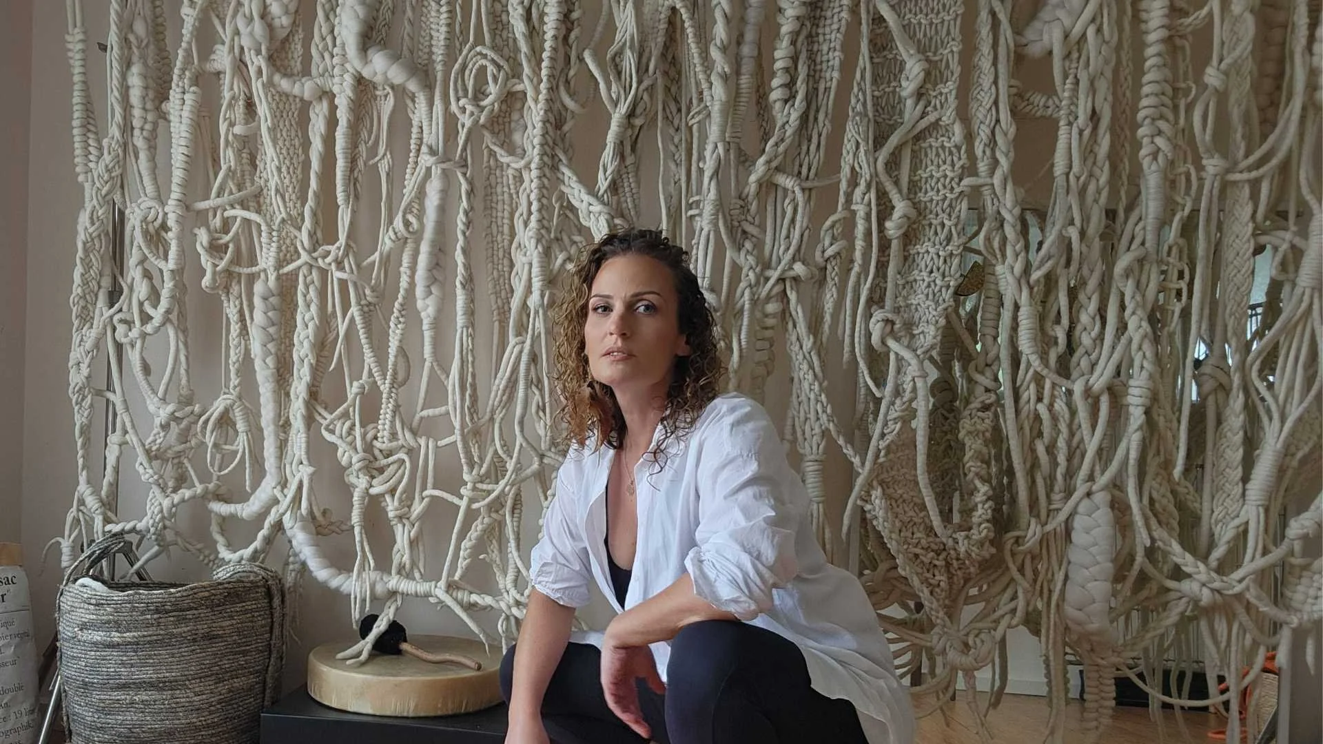 A woman with curly hair and a white shirt posing indoors in front of a large, intricate wall hanging made of knotted, braided and woven off-white cords. There is a woven basket, a small round table with a beige paper bag, and a ruler nearby.