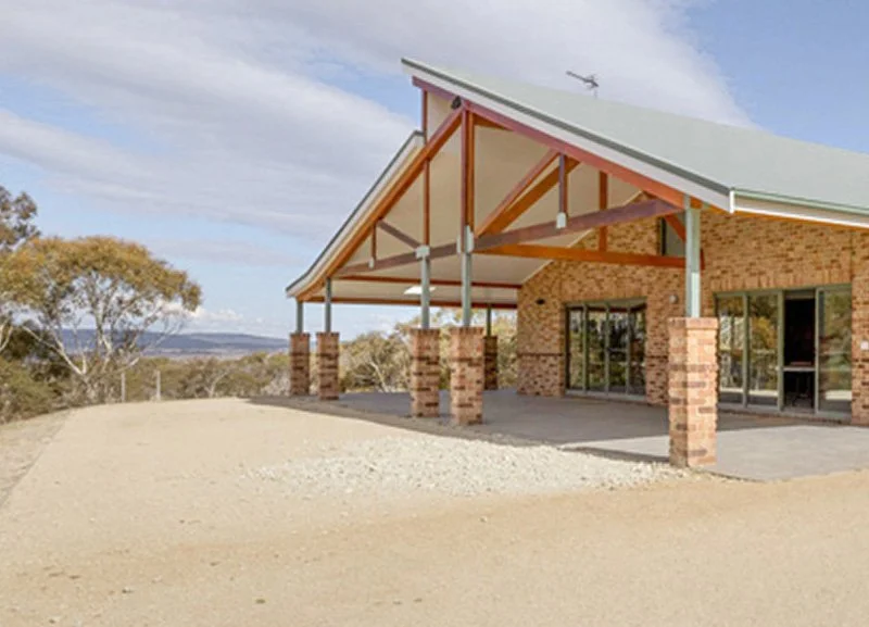 A building with a brick exterior and a large covered porch area supported by brick columns and wood beams, with sliding glass doors, situated in a rural or semi-rural landscape under a partly cloudy sky.