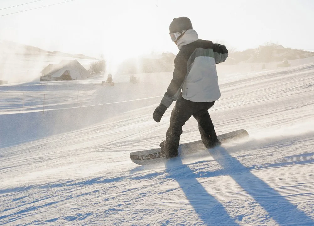 A person snowboarding on a snowy slope in winter, wearing a black and gray jacket, dark pants, gloves, and goggles with the sun shining brightly.