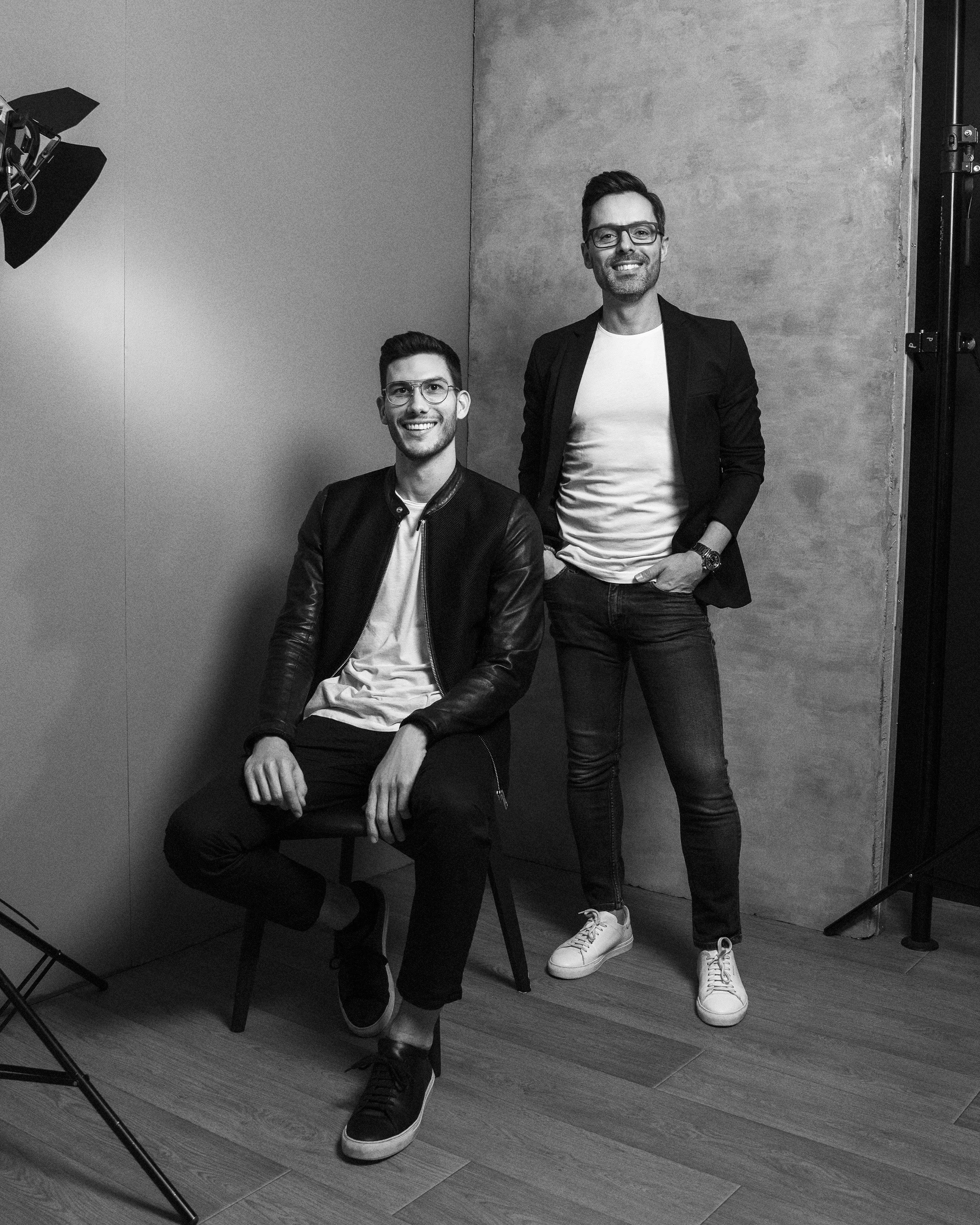 Two young men with glasses posing for a photo in a studio, one seated on a chair and the other standing, both smiling, with a plain wall background, black and white photography.
