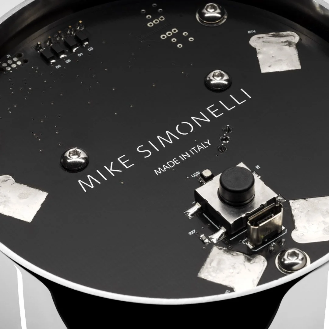 Close-up of the back of a Mike Simoneelli designer alarm clock, showing the brand name and the phrase 'Made in Italy.' The clock features screws, electronic components, and a display area on a black face.