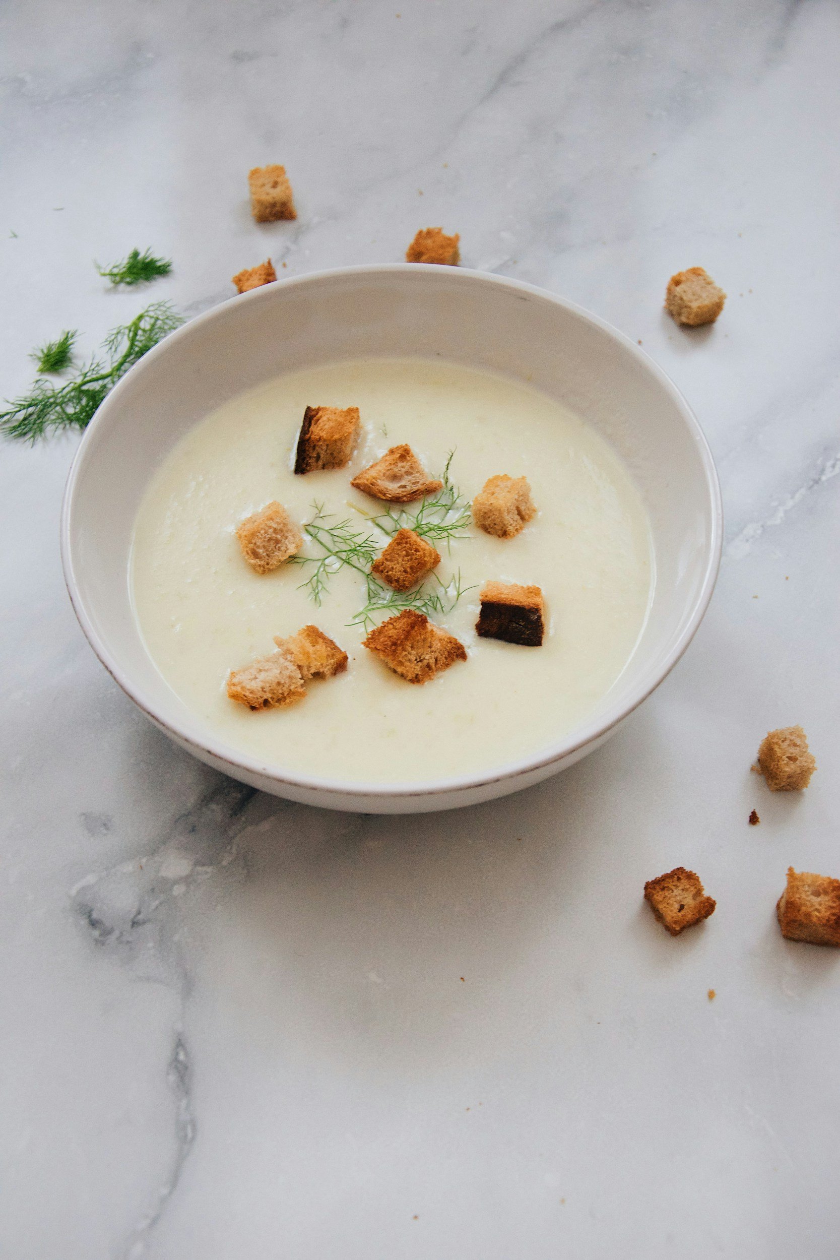 Pale creamy soup in a white bowl topped with golden croutons and green herbs
