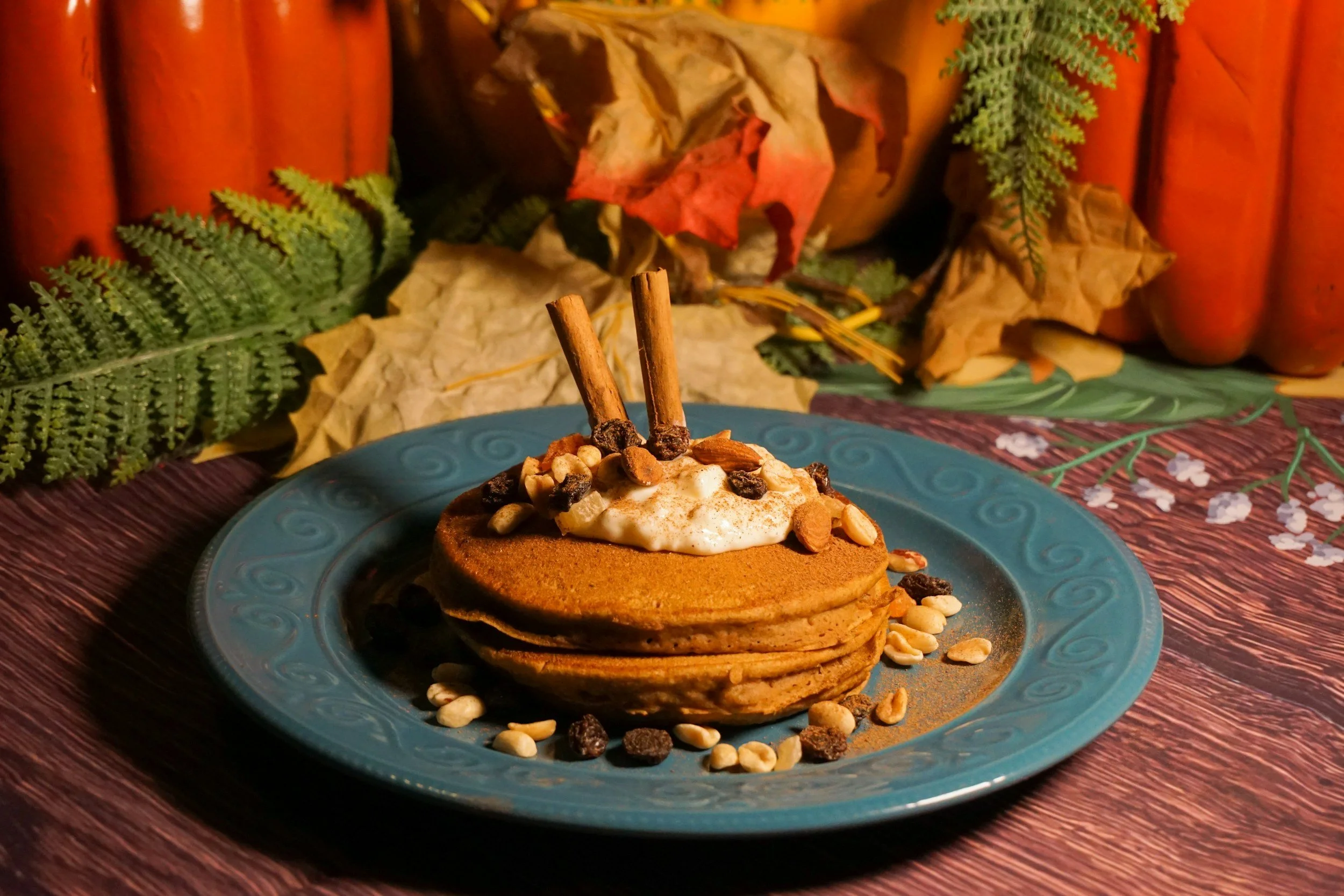 A stack of golden pumpkin pancakes with a creamy topping, nuts and cinnamon sticks