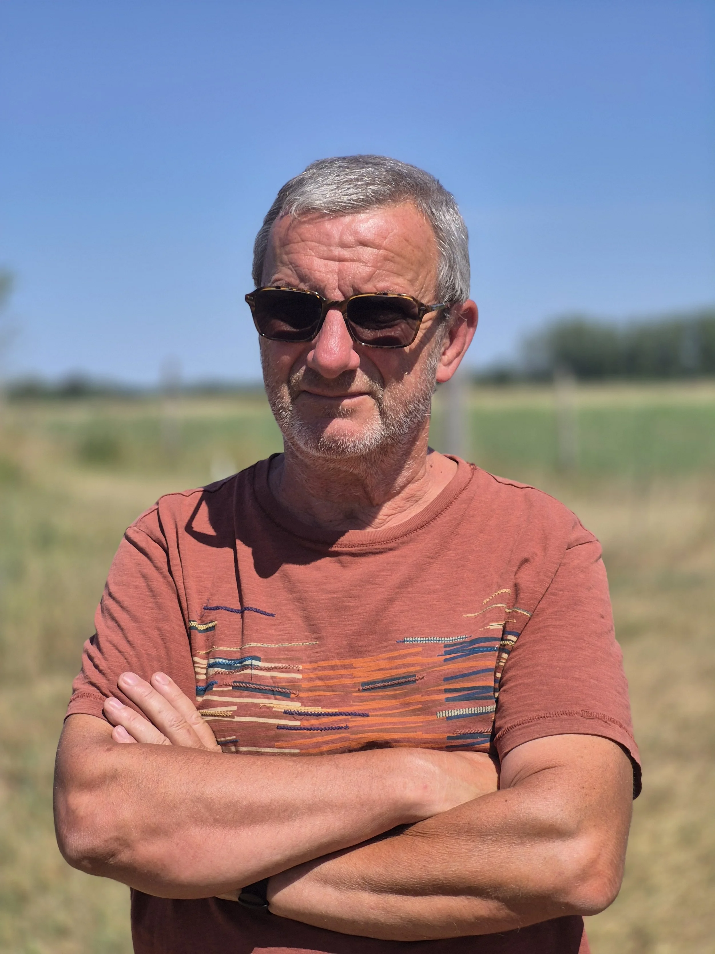 A man with gray hair and beard wearing sunglasses and a red t-shirt with a landscape print, standing outdoors with a blue sky and greenery in the background, crossing his arms.