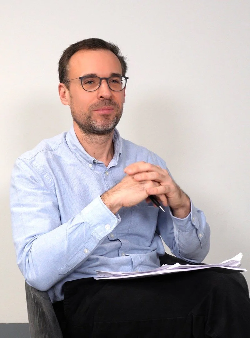 Mann mit Brille sitzt vor weißer Wand, trägt hellblaue Hemd, hält einen Stift in den Händen, und hat Papiere auf dem Schoß.