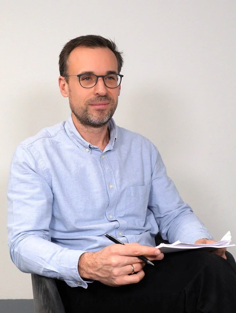 Ein Mann mit Brille sitzt an einem Tisch, hält Stift und Notizblätter, vor einem weißen Hintergrund.