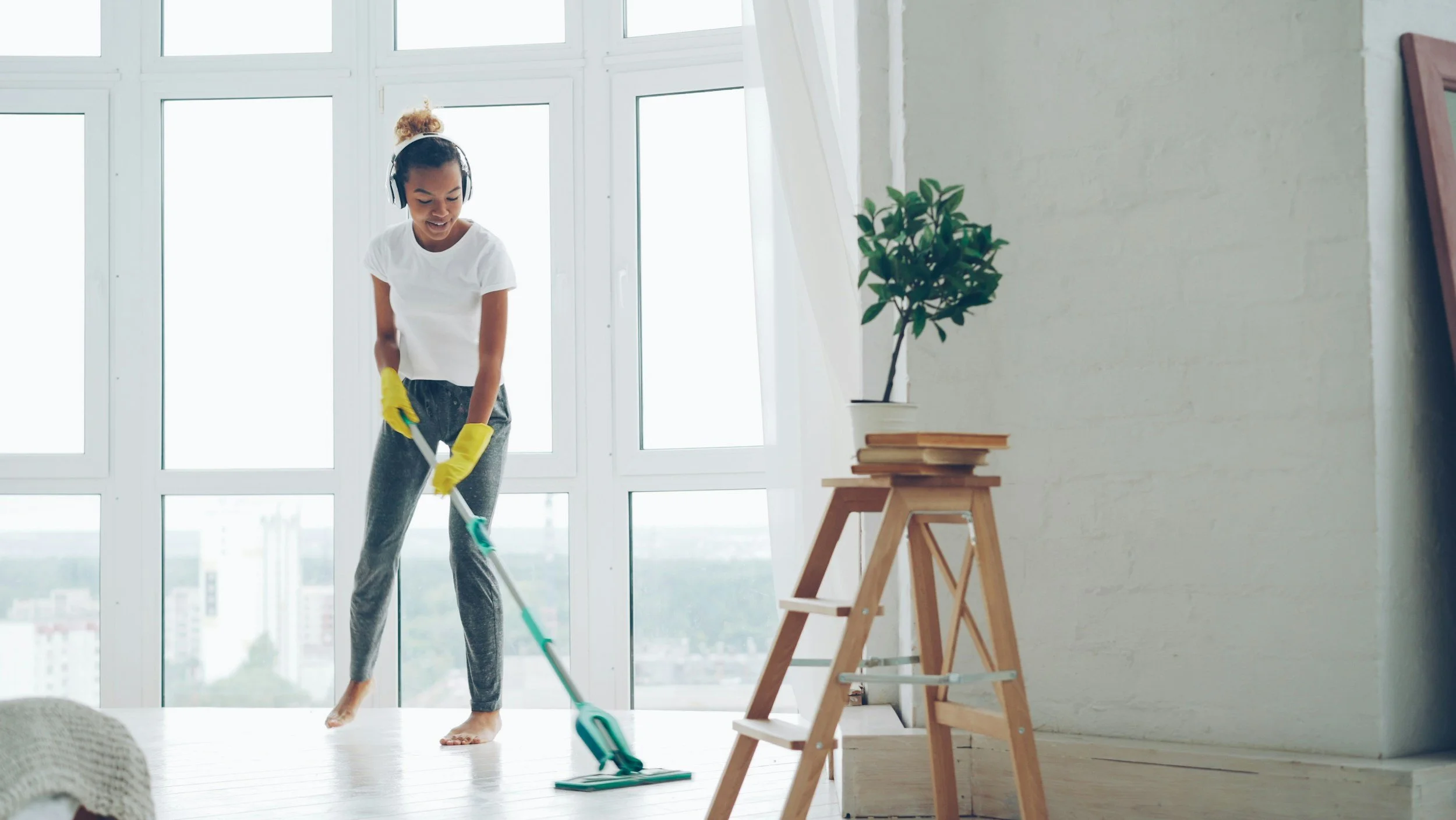 A woman wearing a white shirt, yellow dish gloves, and dark pants. She is mopping the floor around a small latter that has a a stack of books and a potted plant.