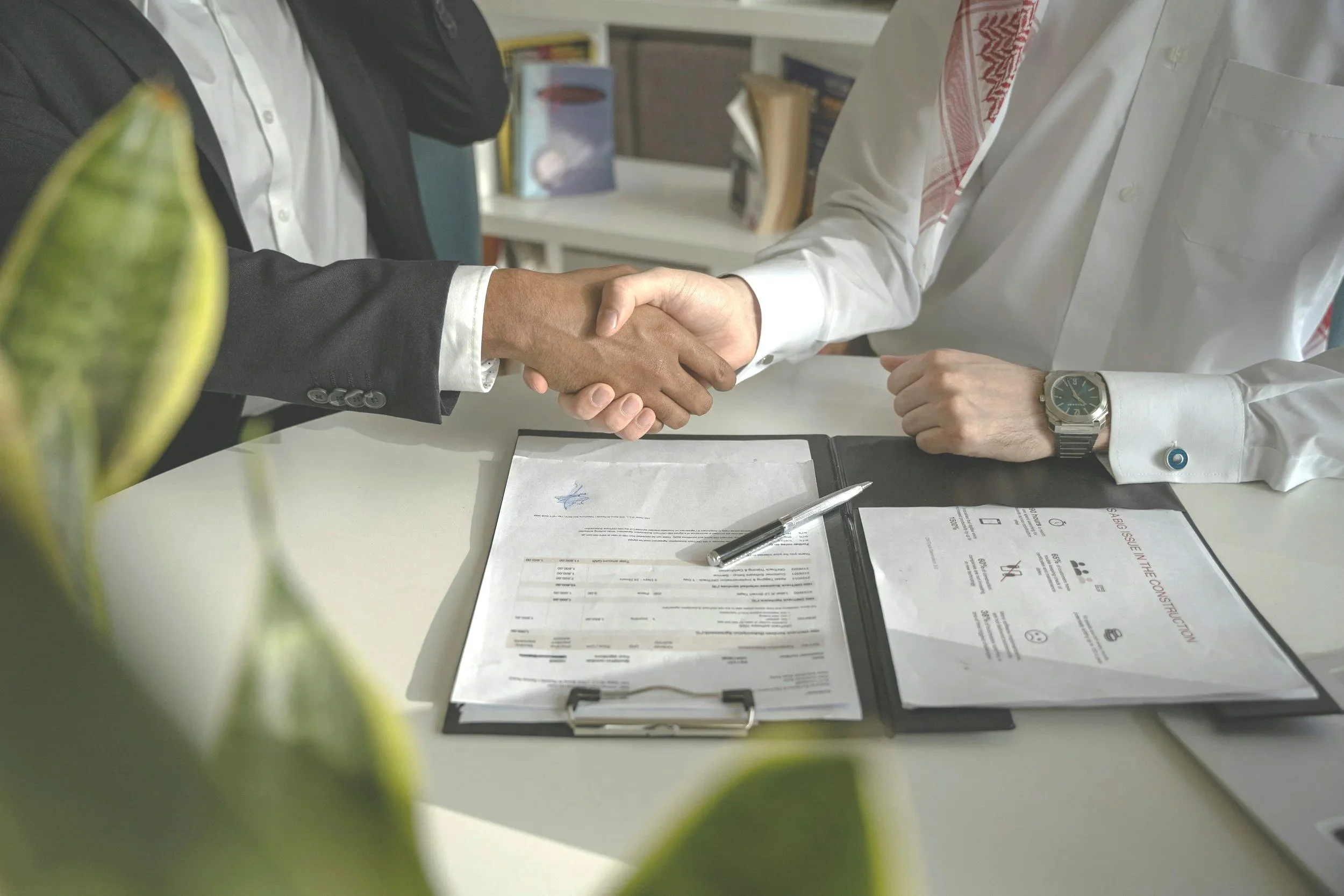 Two men in business attire shaking hands across a desk with documents, a pen, and a clipboard, in an office setting. The foreground shows a blurred green plant.