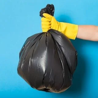 A person wearing a yellow glove holding a black trash bag against a blue background.