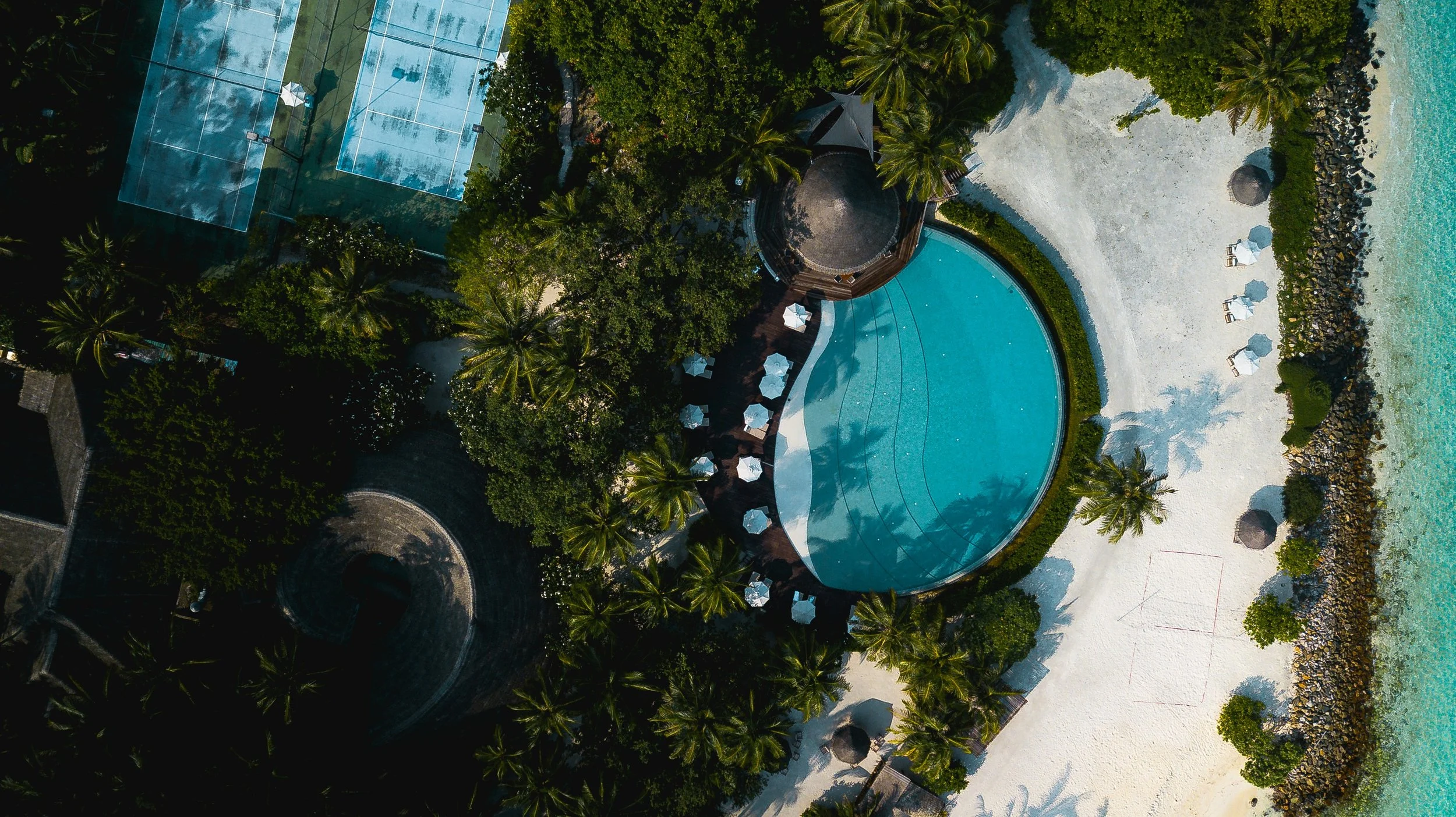 Aerial view of a resort with a swimming pool, surrounded by palm trees and a sandy beach, adjacent to the ocean. Tennis courts are visible to the left, and beach chairs with umbrellas are arranged on the sand.