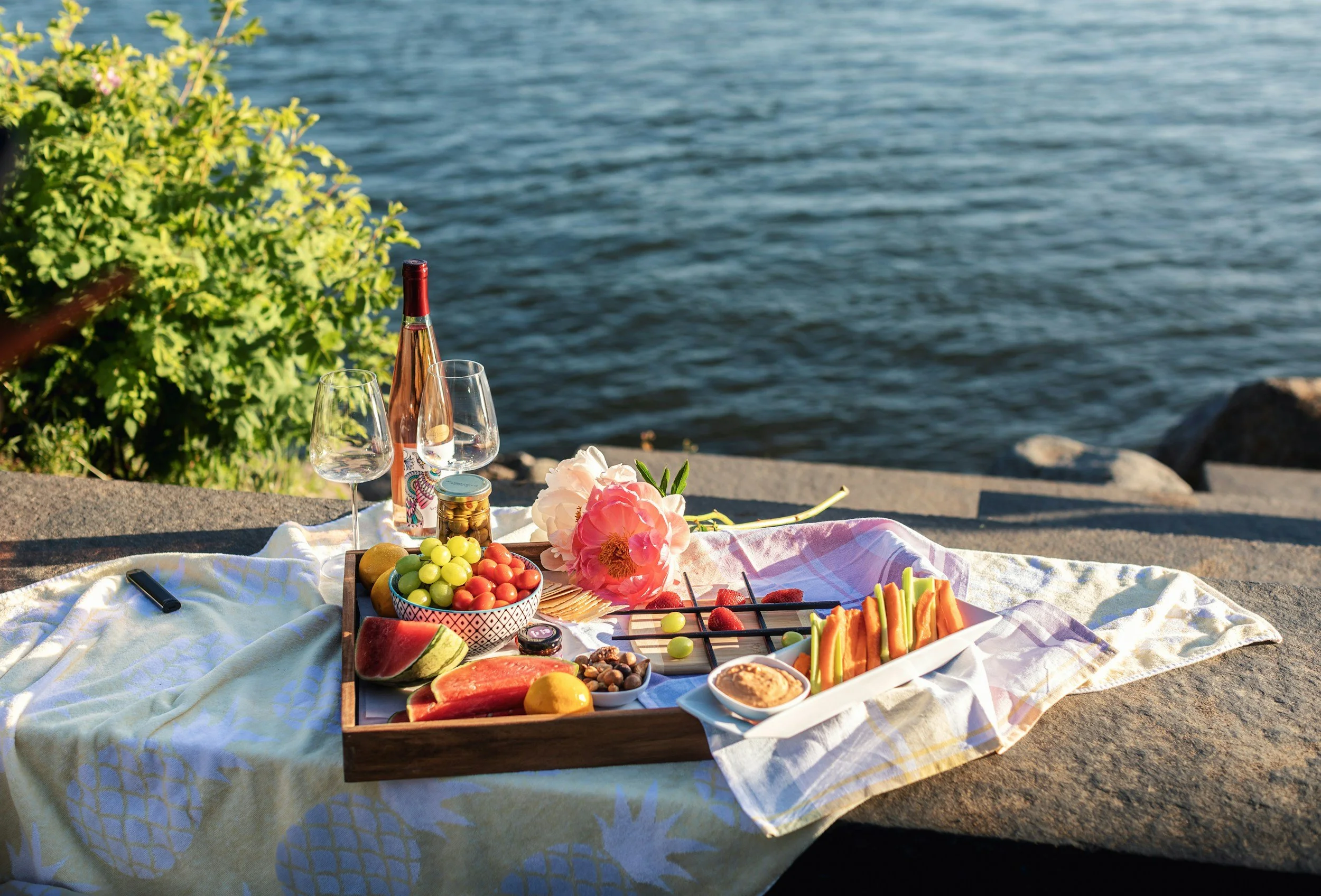 A picnic setup with a tray of snacks and drinks on a blanket by the water, featuring wine glasses, a bottle of rosé, a bowl of fruit, flowers, and assorted vegetables.