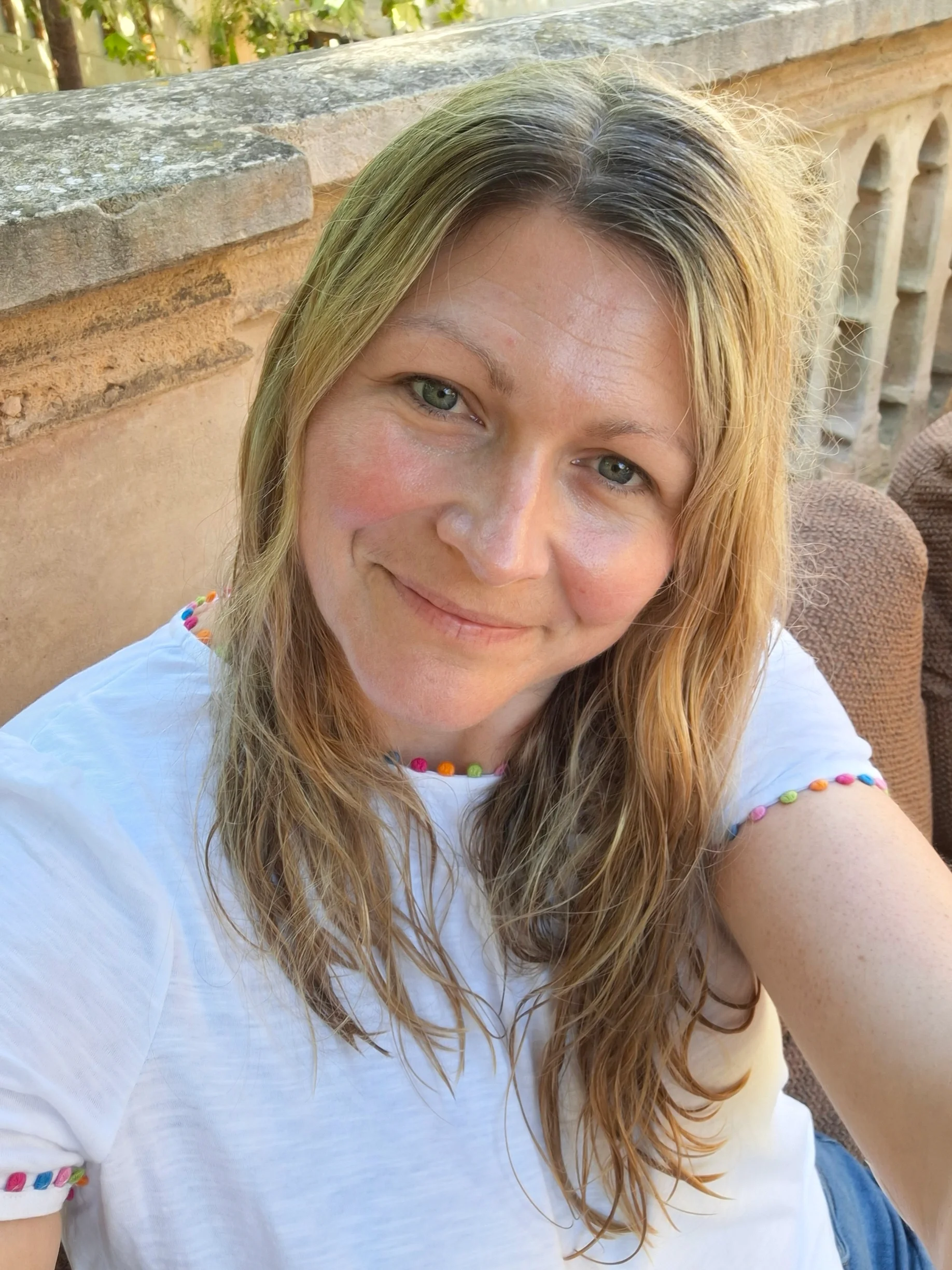 A woman with wavy blonde hair and green eyes taking a selfie outdoors, smiling at the camera, wearing a white shirt with colorful bead details on the sleeves.