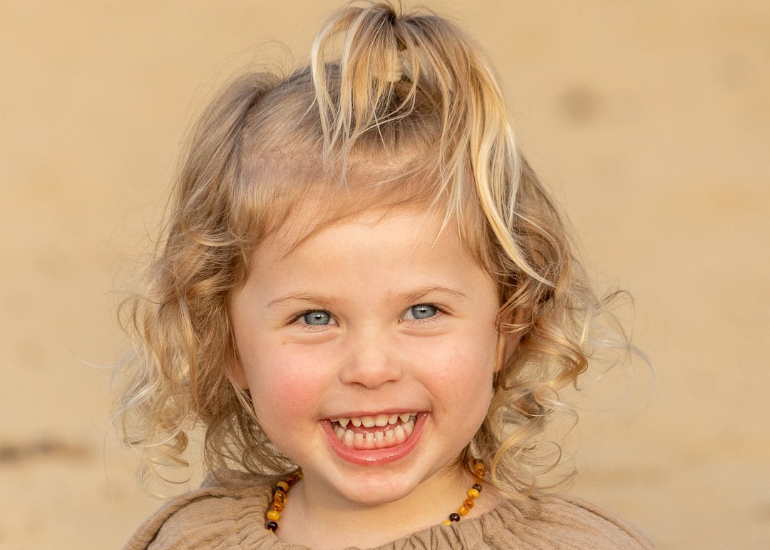 Close-up van een lachend, jong, meisje met blonde, gekrulde haren en blauwe ogen. Zij draag een beige top shirt met een ketting, achtergrond is neutraal en licht, kinderfotografie