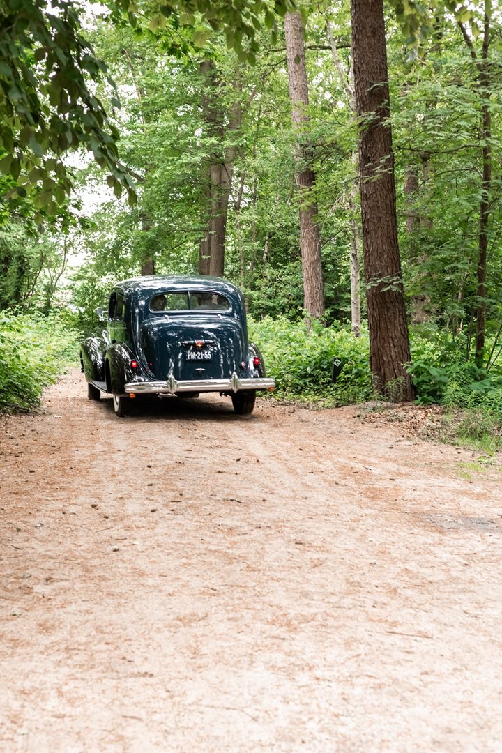 Klassieke trouwauto rijdt, met het bruidspaar weg uit het bos met groene bomen en struiken aan de zijkanten.