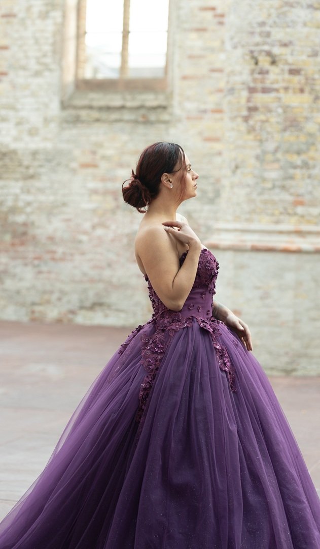 Vrouw in een paarse avondjurk poseert binnen in een prachtige kerk, met een ruwe bakstenen muur en een groot, hoog raam in de achtergrond.