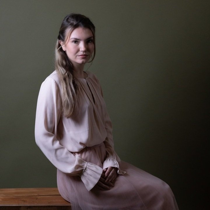 Een jonge vrouw met bruin haar zit op een houten bank, gekleed in een lichtroze jurk, tegen een effen, donkergroene achtergrond, fine art fotografie